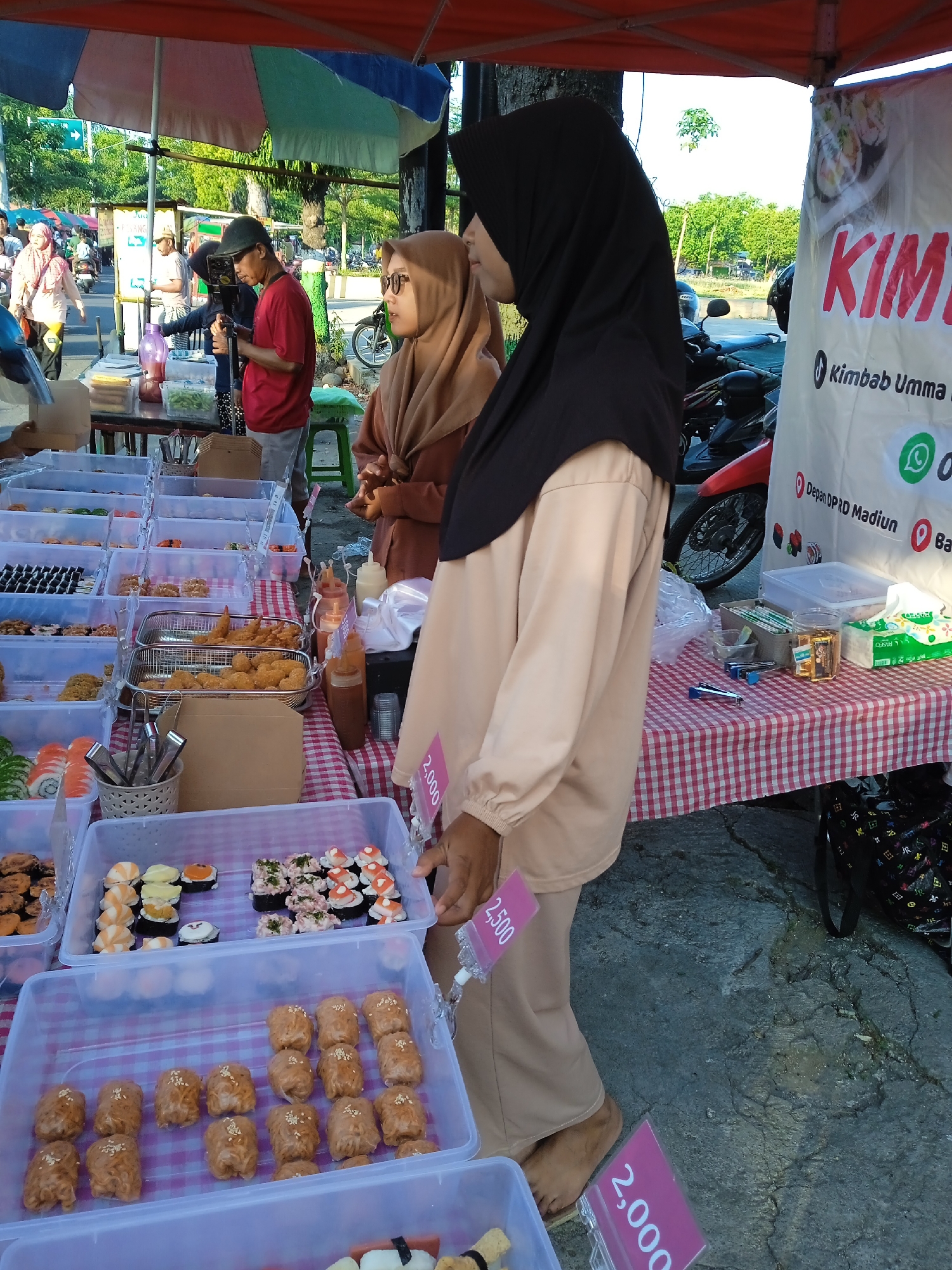 Here is a detailed description of the image:
**Scene:** A street food stall is set up on a sidewalk in what appears to be Madiun, Indonesia. The stall is under a red canopy. The stall sells Kimbab Umma, a Korean-style sushi.
**People:** Two women wearing hijabs stand behind the stall, presumably running the business. The woman closest to the camera wears a black hijab and beige clothing. A second woman behind her wears a brown hijab and coordinating clothing. A few other individuals are seen in the background, including a man wearing a red shirt and baseball cap, looking at a phone.
**Objects:** The stall has multiple clear plastic containers filled with various types of sushi. There are price tags next to some containers, indicating the cost of the sushi. A red and white checkered tablecloth covers the table. There are bottles of sauce, napkins, and other supplies on the table. A sign next to the stall promotes "Kimbab Umma," including contact information and a location "Depan OPRD Madiun." Motorcycles are parked behind the stall.
**Actions/Interactions:** The two women are standing behind the stall, possibly waiting for customers. Customers are browsing the sushi selection.
**Environment:** The scene is set outdoors on a sunny day. Trees and buildings can be seen in the background, indicating an urban area. The ground is paved.