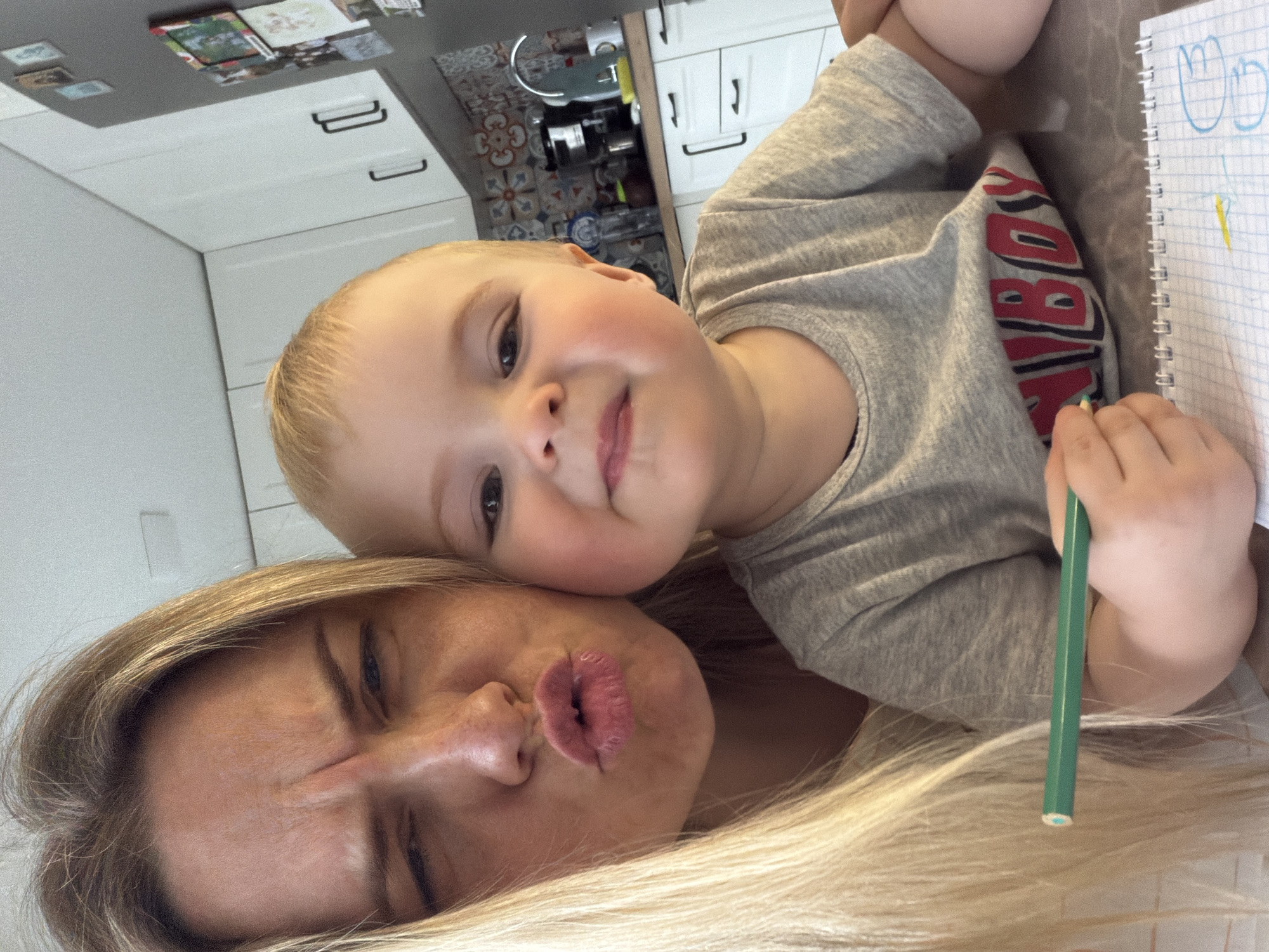 A selfie shows a woman and a young child in what appears to be a domestic setting, possibly a kitchen. The woman, with blonde hair, is making a kissing face towards the camera. The child, with blonde hair and wearing a grey t-shirt with red lettering, is looking at the camera and holding a green pencil. A notebook is visible on the right side of the frame. The background shows kitchen cabinets, a countertop, and a tiled backsplash. There is no evidence of the image being taken in the city of Votkinsk, Russia.