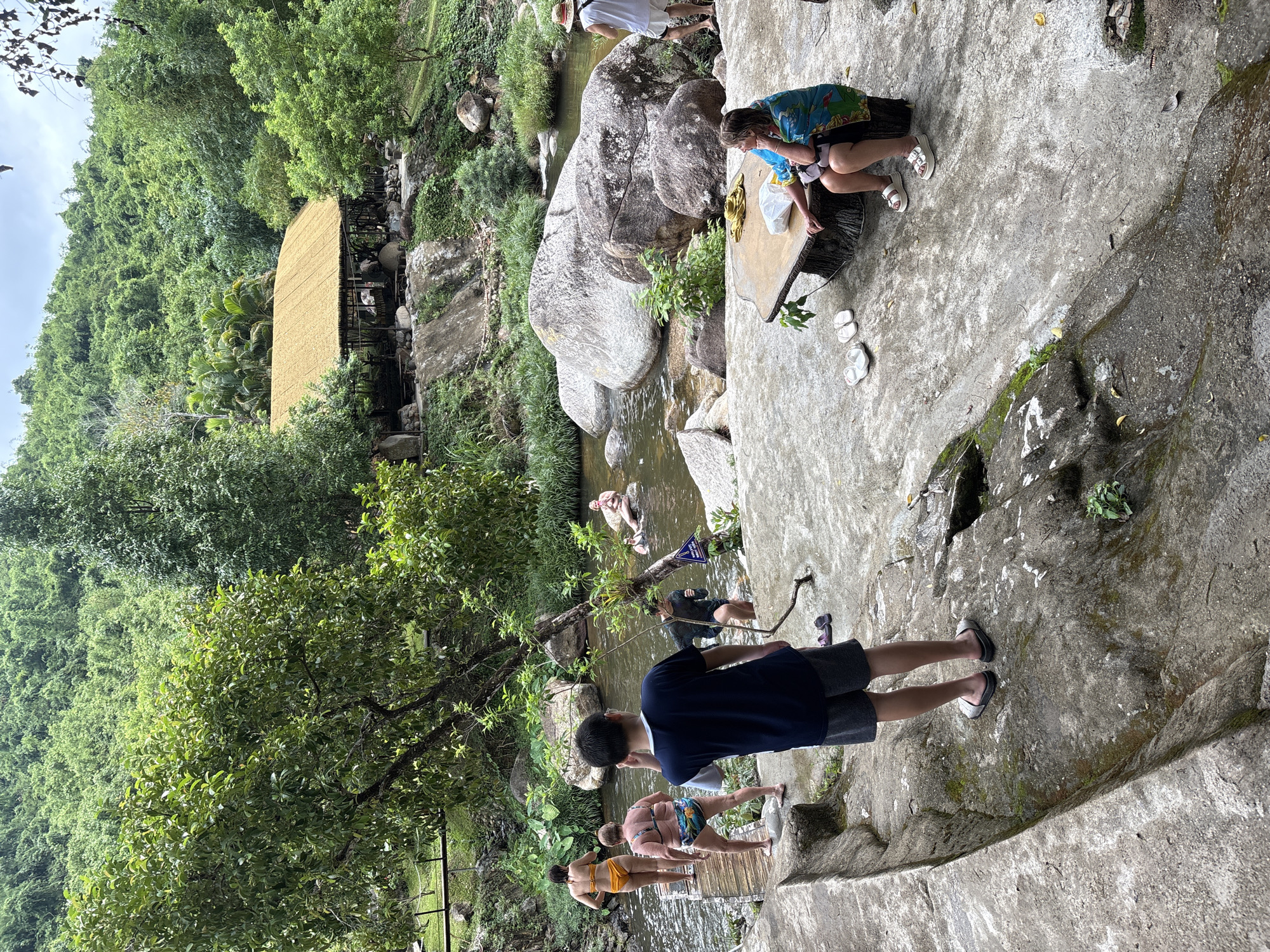 This is a photograph of a resort near Khanh Vinh, Vietnam. The resort features a river with a sandy bottom and large rocks, surrounded by lush greenery and a bamboo-roofed building. Several people are visible in the image. In the foreground, a young person wearing a dark blue t-shirt and grey shorts stands facing away from the camera, looking towards the river. To the left of the river, a wooden walkway leads to a group of people. One woman in an orange bikini and another woman in a patterned swimsuit are walking on the walkway. Further into the river, several individuals are swimming or wading. On the right side of the frame, a woman in a blue and green sarong is seated at a rustic wooden table, with another person in a white shirt and shorts standing nearby. The environment appears to be a natural outdoor setting with a man-made resort infrastructure.