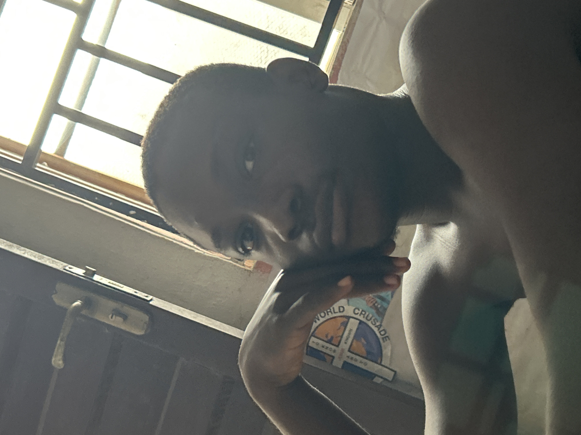 A young person with dark skin and a close-cropped haircut is positioned in the foreground, their face turned to the left, resting their chin on their left hand. The background features a closed door with a metallic handle and lock on the left, and a barred window with bright light filtering through on the right. A portion of a white T-shirt with a circular logo is visible on the person's chest. The lighting suggests an indoor setting, possibly in a residence, in Ihiagwa, Nigeria, given the overall ambiance and the presence of features common to Nigerian homes.