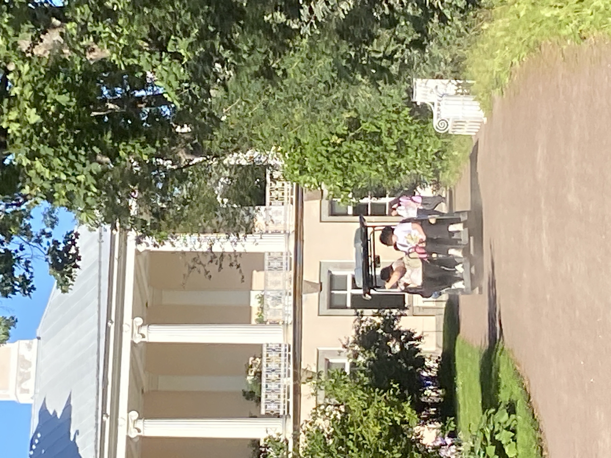 This image depicts a sunny day scene at the Catherine Palace in Pushkin, Russia. In the foreground, a dirt path leads toward a light-colored building with classical architectural elements, including large columns. A small electric vehicle, resembling a golf cart, is driving away from the viewer down the path. It carries at least three people. Two individuals are visible in the front seats, and a third person is in the back. The person in the front left appears to be wearing a light-colored top and dark pants. The person in the front right is wearing a white t-shirt and dark pants. A third person, seen from behind in the back, is wearing a pink jacket. To the left of the path, there is a well-maintained lawn with some green bushes and flowering plants. To the right of the path, lush green trees and bushes line the edge. The building in the background has a light-colored facade and a prominent balcony with ornate railings. The sky above is a clear blue. The overall impression is one of a peaceful and scenic park setting within a historic estate.
