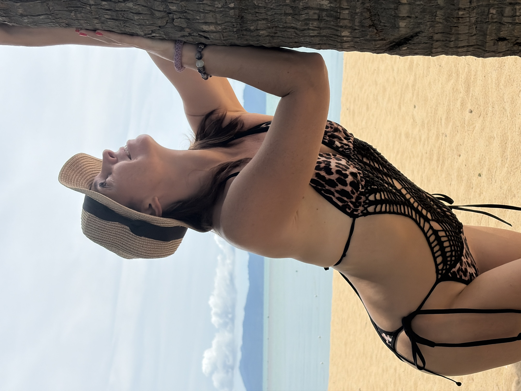 In Baan Bo Phut, Thailand, a woman wearing a leopard-print bikini and a straw hat leans against a palm tree on a sandy beach. Her right hand is placed on the tree, and her left arm is bent, with her hand near her chest. She is looking upwards with a slight smile. The ocean is visible in the background, with a hazy mountain range on the horizon. The sky is bright blue with scattered clouds.