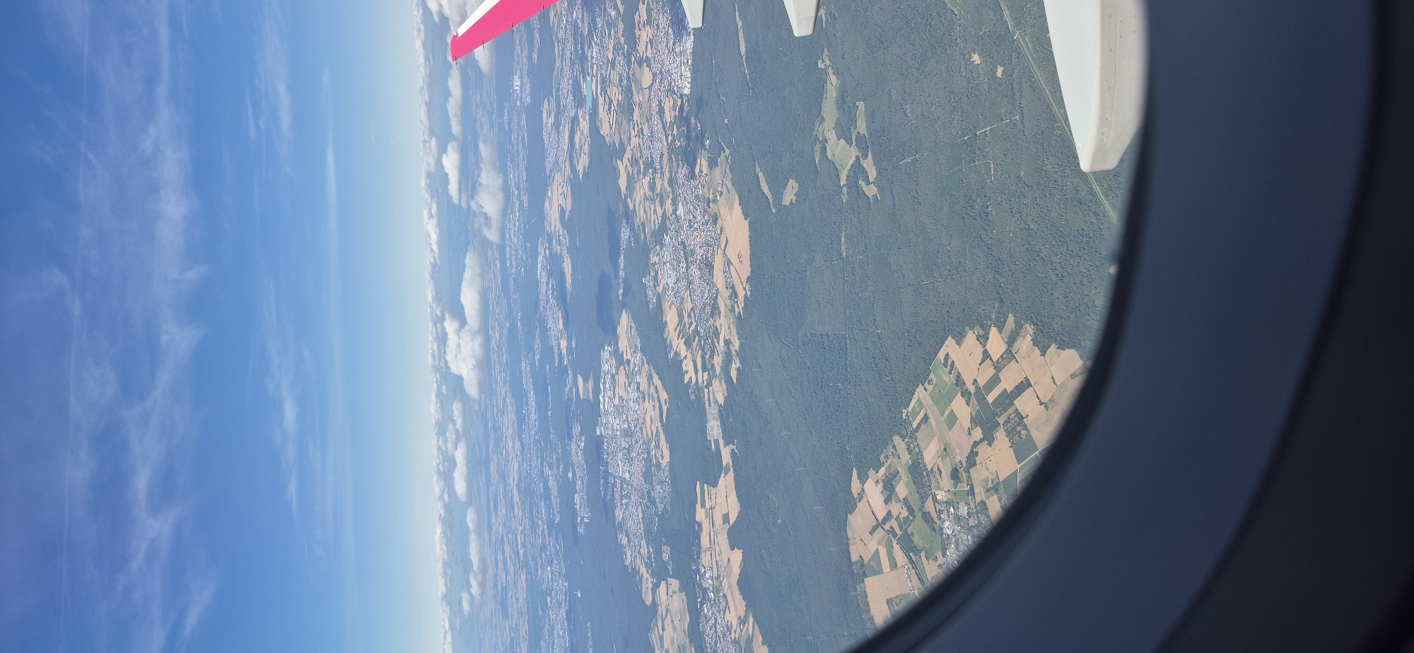 An aerial view from an airplane window shows a landscape dotted with agricultural fields, forests, and urban areas. The sky above is a bright blue with scattered white clouds. The airplane wing, with its pink tip, is visible on the right side of the frame. The ground below consists of a patchwork of green forests and lighter colored agricultural fields, some appearing to be recently harvested. Small towns and clusters of buildings are interspersed within the landscape. The perspective is from a high altitude, looking down at the earth. The location is identified as Spata-Loutsa Municipal Unit, Greece.
