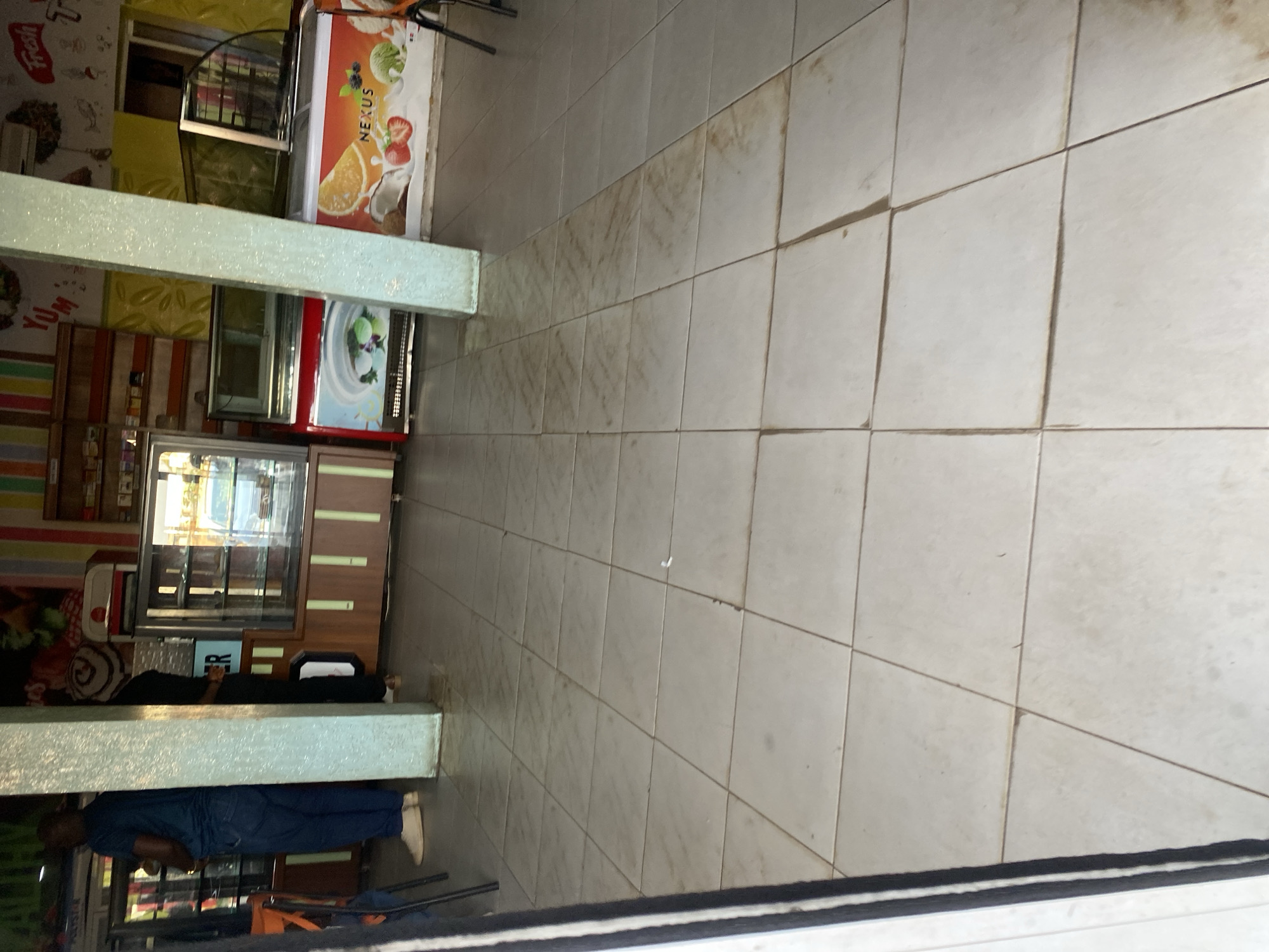 The image depicts the interior of an establishment, possibly a cafe or restaurant, with tiled flooring. In the background, there are display cases containing food items, likely pastries or ice cream, and refrigeration units. One refrigeration unit prominently features a "NEXUS" logo with an illustration of fruits and ice cream. A person is visible in the left of the frame, wearing a blue shirt and dark pants, standing near a counter. Another person is partially visible behind a pillar. The overall environment appears to be a commercial space in Barakin Lerawi, Nigeria, with decorative elements on the walls and shelves displaying products. The lighting suggests an indoor setting.