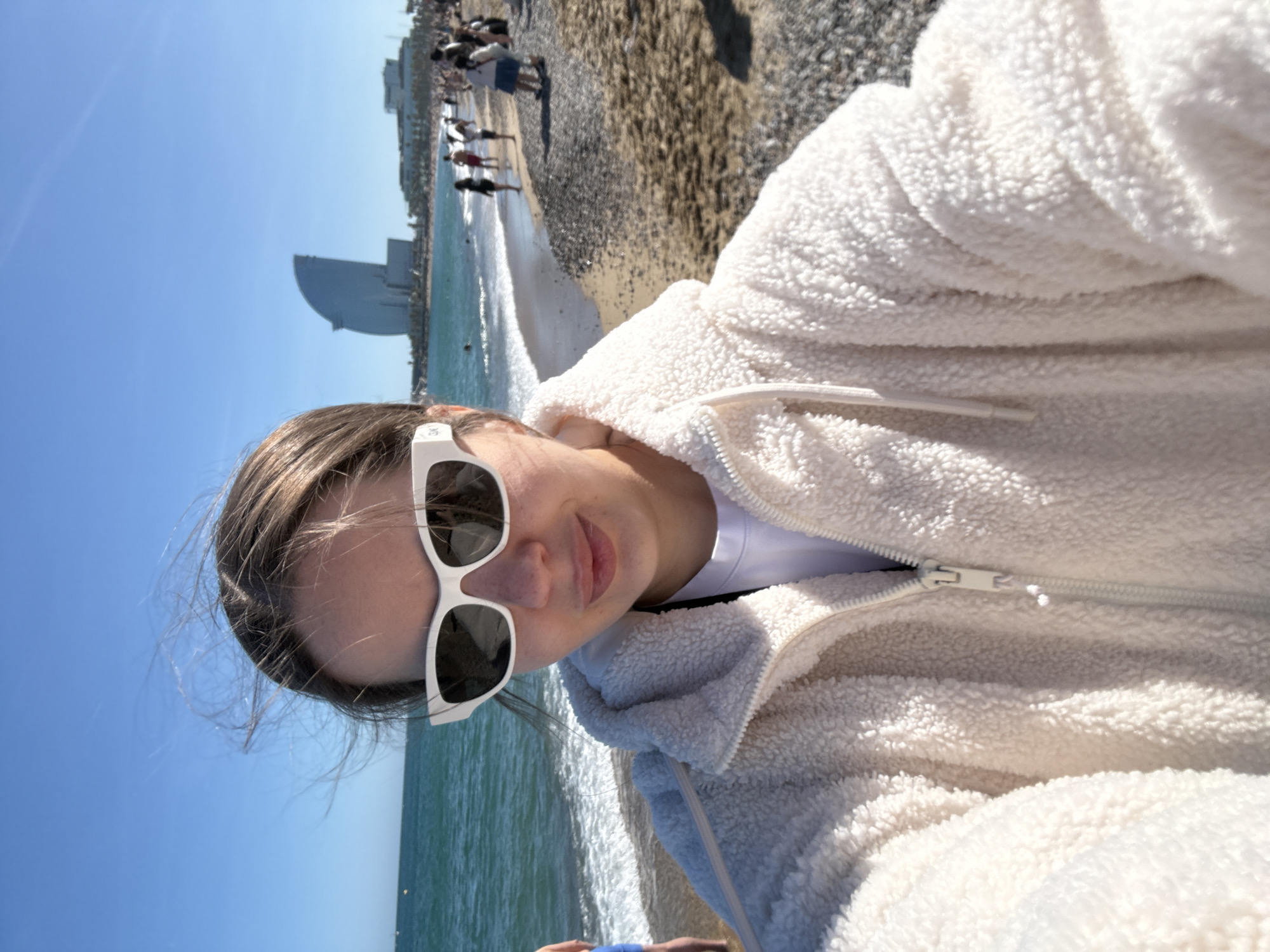 A woman with light brown hair and sunglasses is taking a selfie at Barceloneta Beach in Barcelona, Spain. She is wearing a white, fleece-like jacket over a white t-shirt. The blue sky and sea are visible in the background. People are walking along the beach in the distance. The Hotel Arts Barcelona, a distinctive skyscraper with a sloping roof, is also visible in the background. The beach is a mix of sand and pebbles. The sun is shining.