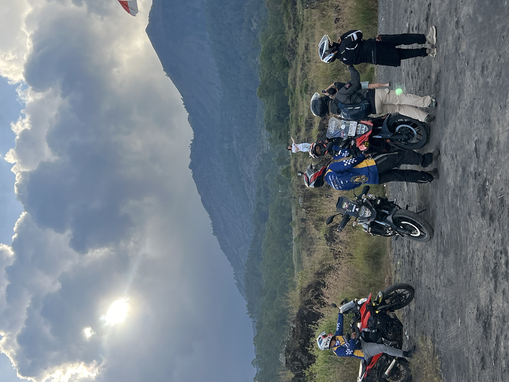 A group of people on motorcycles has stopped on a paved surface. There are four visible motorcycles; the closest one is red and black, the next is black, and the remaining two are partially obscured. The riders are wearing helmets and riding gear. One person is standing without a motorcycle. The surrounding environment is a hillside with sparse vegetation and a large mountain in the background, beneath a cloudy sky with the sun visible behind the clouds. The location is Kintamani, Indonesia.