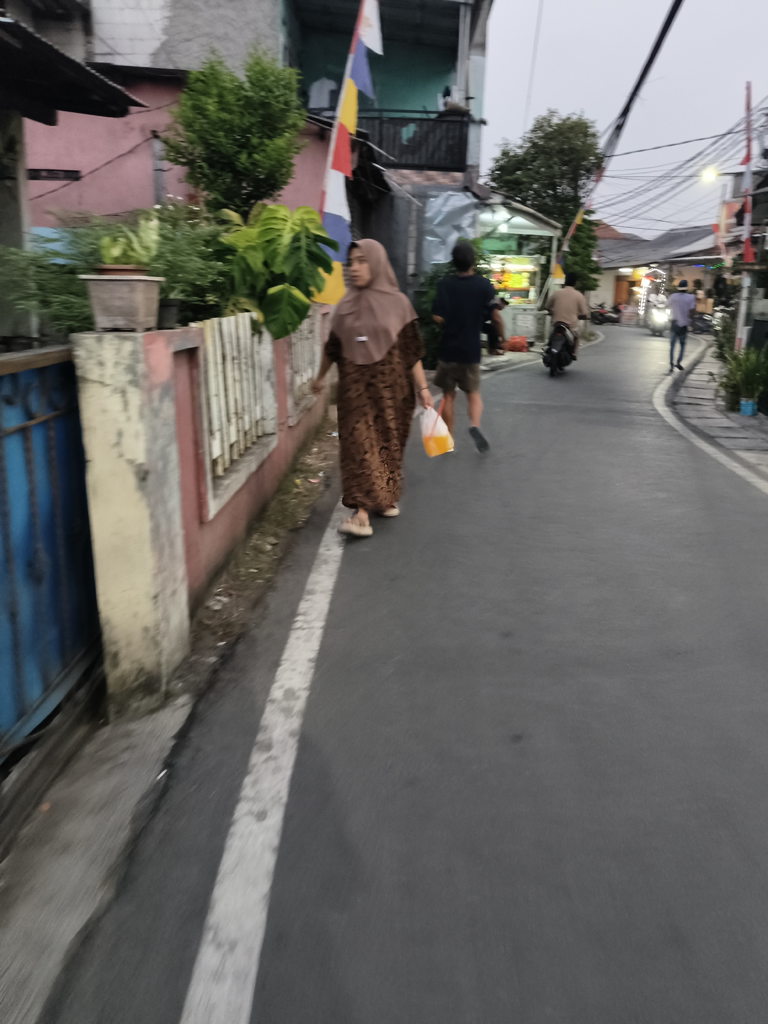 The image depicts a street scene in a residential area, likely in South Tangerang, Indonesia, given the presence of typical Indonesian urban infrastructure and architecture. The time of day appears to be late afternoon or early evening, with diffused light.
In the foreground, a woman wearing a brown hijab and a patterned brown dress is walking away from the viewer, holding a transparent plastic bag with an orange liquid inside. Her feet are clad in light-colored sandals. To her left, a weathered fence borders a property with a pink wall and some greenery.
Further down the street, other individuals are visible. A person in a dark shirt and shorts is walking ahead of the woman, also moving away from the camera. Two motorcycles are on the street; one is ridden by a person wearing a light brown shirt and dark shorts, and another is parked on the side of the road. Another individual in a blue shirt and dark pants is walking towards the foreground on the right side of the street. The street itself is paved and has a white line marking the edge. Buildings with multiple stories line the street, some with balconies and electrical wires overhead. A small food stall or kiosk with lights is visible in the mid-ground. The overall environment suggests a bustling, ordinary day in an Indonesian neighborhood.