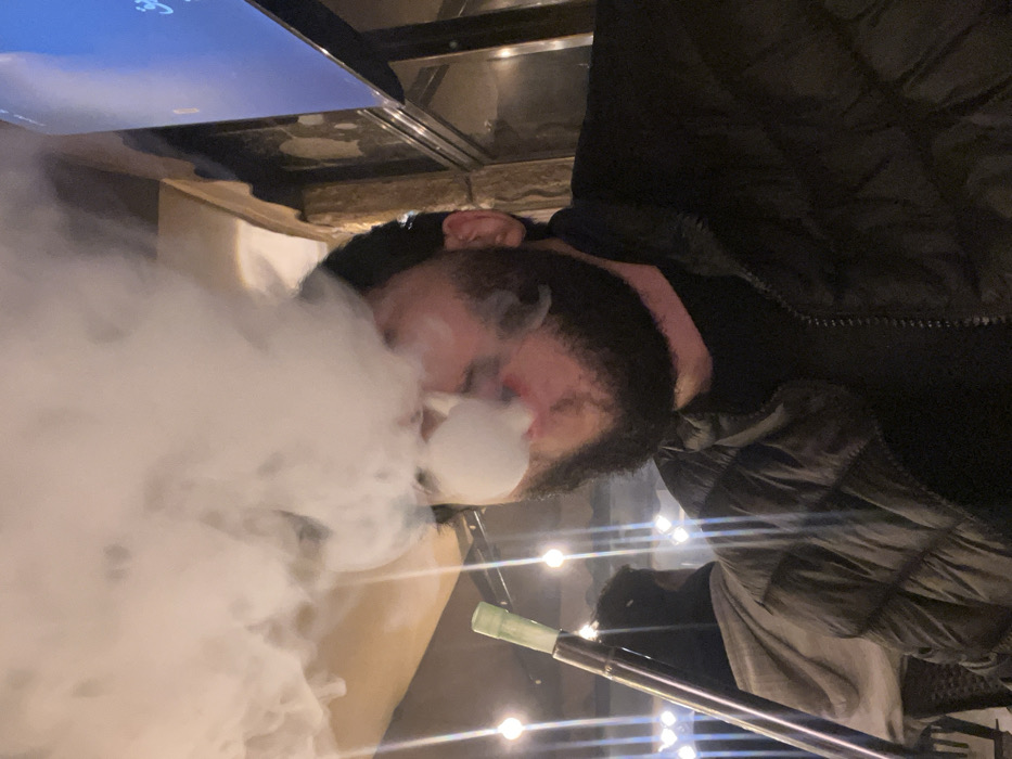 A person wearing a dark jacket exhales a large cloud of smoke, presumably from a hookah. The person has dark hair and fair skin. A hookah hose with a green tip is visible. The setting appears to be indoors with several lights. A screen or television is partially visible in the upper left corner. The image is rotated.