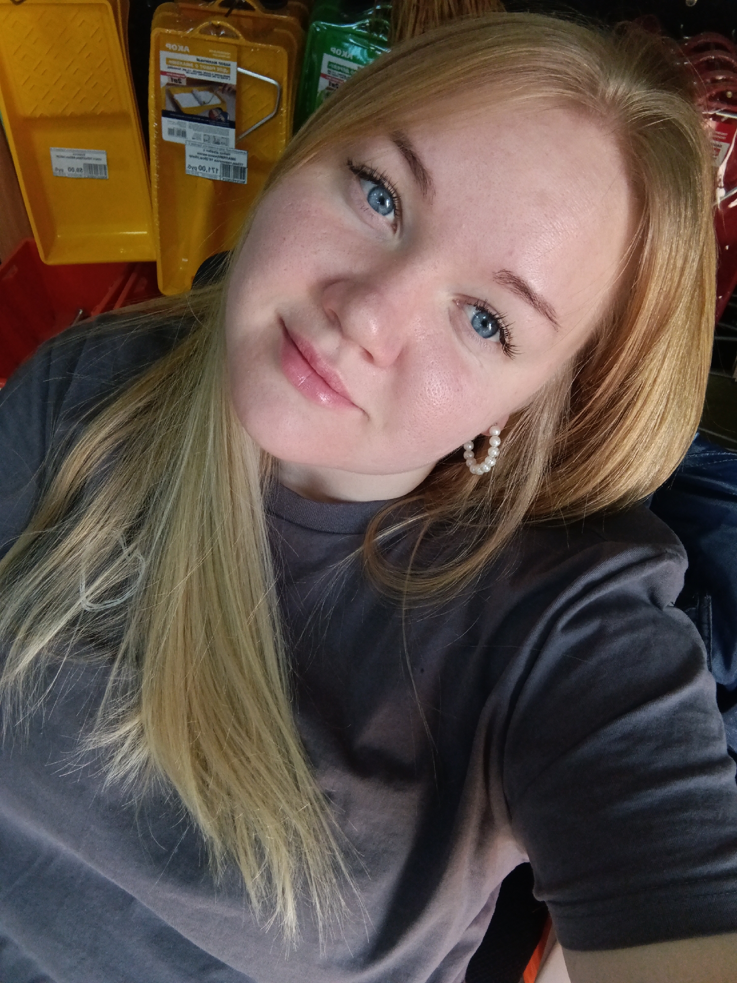 A young woman with blonde hair and blue eyes is taking a selfie. She is wearing a gray t-shirt and pearl earrings. Behind her are shelves with painting supplies, including paint trays and rollers. The location is Unknown, Unknown.