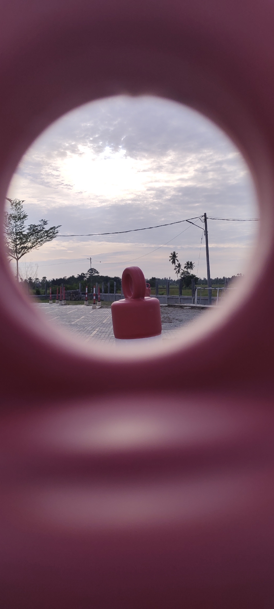 The image shows a red, bottle-shaped object with a loop on top, centered in the foreground, viewed through a circular opening of a larger red, cylindrical structure. The object rests on a paved surface. Beyond the object, through the circular opening, is a landscape featuring a flat area with a fence, poles alternating red and white, trees including palm trees, and a utility pole with wires. The sky is overcast. The location is Meunasahaluedrienlhoksukon Barat, Indonesia.