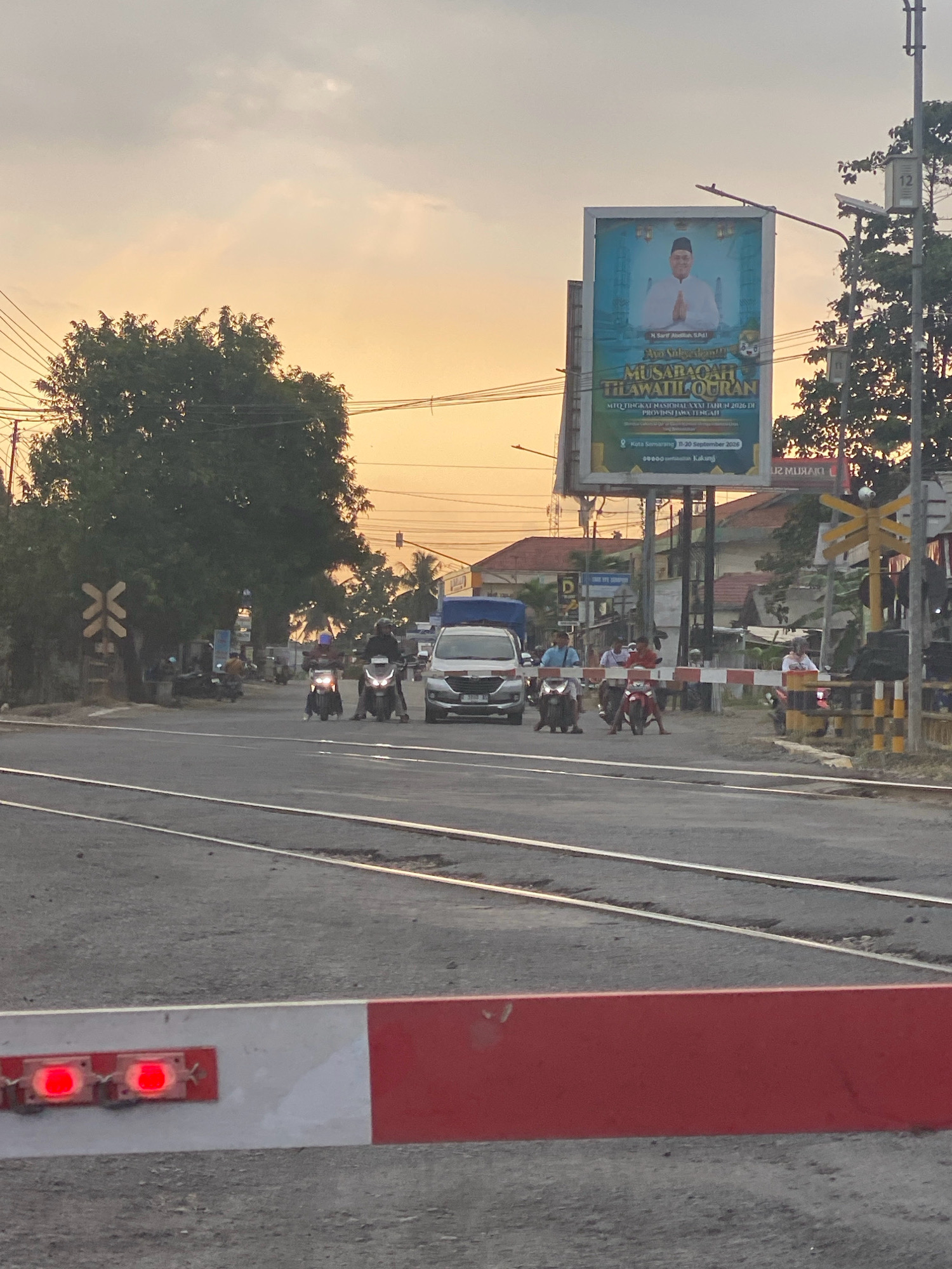 This image captures a view of a railway crossing in Sumpiuh, Indonesia, during sunset. The sky is painted with warm hues of orange and yellow, indicating the approaching evening. A prominent billboard stands on the right side of the frame, displaying an image of a man in religious attire, likely a cleric, with text below in Indonesian. The billboard advertises an event related to the recitation of the Quran.
In the foreground, a red and white railway crossing barrier is lowered, with two red lights flashing to warn of an approaching train. Several motorcycles are stopped behind the barrier, waiting for it to be raised. A white van is also visible, positioned behind the motorcycles. The railway tracks are clearly laid out across the road.
On the left side of the image, a large tree with dense foliage stands tall. Further in the background, several buildings and other trees can be seen, suggesting a suburban or semi-urban environment. Power lines crisscross the sky, typical of many Indonesian cityscapes. The overall scene depicts a moment of stillness and anticipation at a busy intersection, awaiting the passage of a train.