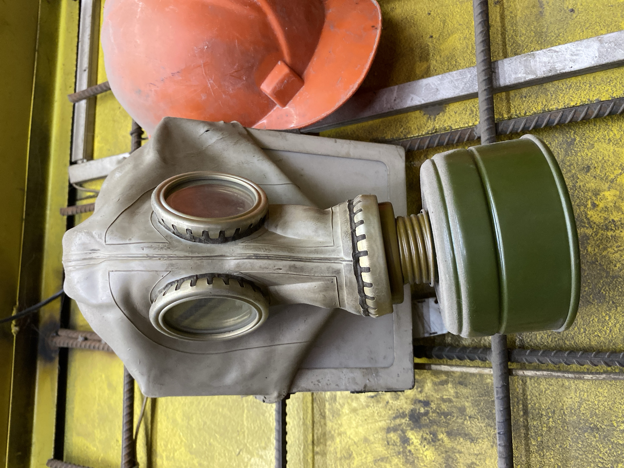 A close-up, eye-level shot shows a worn, light gray gas mask hanging against a yellow, textured surface. The gas mask has two round, glass eyepieces with metallic rims and a green, cylindrical filter attached to the bottom. To the right of the gas mask, partially visible, is an orange hard hat. The objects appear to be resting against or hanging on a structure made of rebar and metal bars. The background is a brightly lit, rough, yellow surface, possibly a wall or a large piece of equipment. The lighting is direct, casting faint shadows. The image does not provide any information about the city of Bishkek, Kyrgyzstan, or any specific location within it. There are no people or discernible actions in the scene.