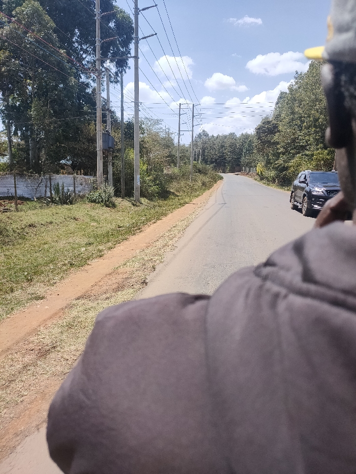 The image captures a roadside scene in Ewuaso Oonkidong'i ward, Kenya, on a bright, sunny day with scattered white clouds in a blue sky. The perspective is from behind a person, whose dark grey or black hooded shoulder and the back of their head with a yellow detail on a cap are visible in the foreground, partially obscuring the lower right of the frame.
A paved asphalt road, seemingly recently constructed or well-maintained, stretches into the distance, curving gently to the left. Along the left side of the road, multiple tall utility poles with numerous power lines run parallel, suggesting an established infrastructure. One pole features a prominent grey transformer box. Below the utility lines, a dense line of trees and various green bushes borders the road. Further left, behind the vegetation, a corrugated metal fence is visible, alongside patches of grass and smaller plants.
On the right side of the road, a dark-colored SUV, possibly a Toyota Fortuner, is visible in the middle distance, driving or parked facing away from the viewer. More dense trees and foliage line this side of the road as well. The ground immediately adjacent to the road is a mix of reddish-brown soil and dry grass. No text is visible in the image.