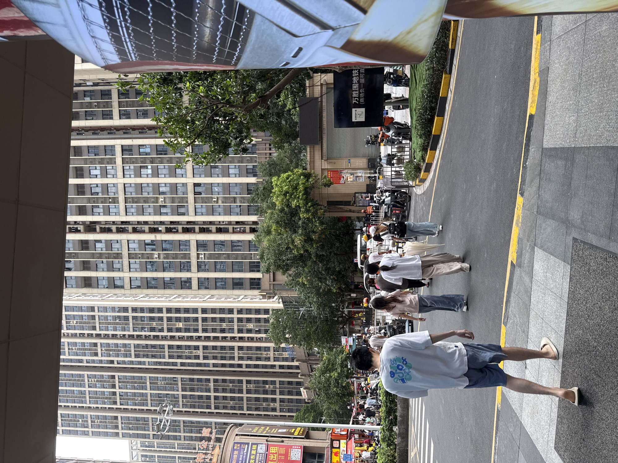 In Haizhu District, China, a street scene unfolds with a focus on pedestrians and urban architecture. The foreground features a person wearing a white t-shirt with a blue circular graphic and light blue shorts, walking away from the camera on a sidewalk. The sidewalk is paved with gray tiles and delineated by dark gray borders. Behind this individual, a crosswalk with white stripes is visible, with several people walking across.
Further back, a group of women are walking towards the right side of the frame. One wears a light pink t-shirt and light blue jeans, another a white t-shirt and beige pants, and a third in a white t-shirt and dark pants. One of the women carries a light-colored umbrella. The street is paved with asphalt and bordered by a curb painted with alternating black and yellow stripes.
In the background, towering apartment buildings with numerous windows dominate the skyline. Trees line the street, providing a touch of greenery. Several motorcycles and scooters are parked along the curb, suggesting a busy urban environment. A black sign with Chinese characters and a white QR code is posted on a building. The overall impression is a snapshot of daily life in a bustling Chinese city.