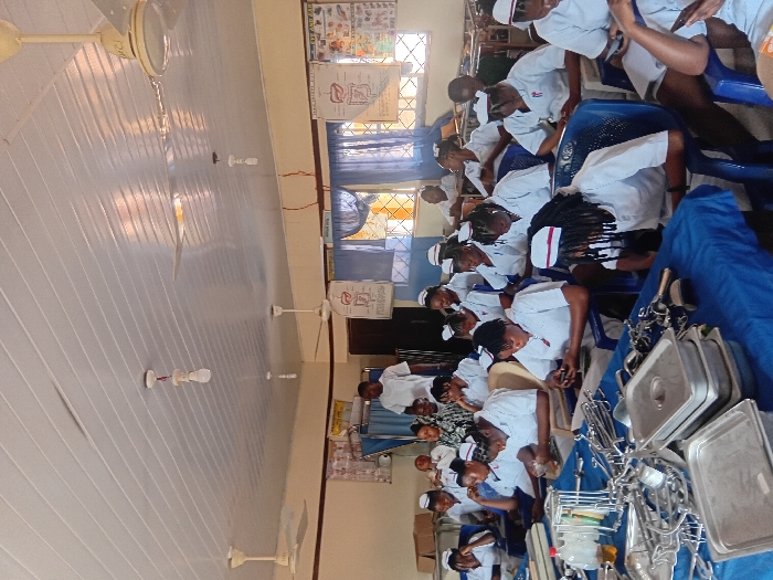 An indoor educational setting, likely a classroom or laboratory, is observed in Ejule, Nigeria. Approximately 25-30 individuals, predominantly young females, are present, dressed in white nursing uniforms with white caps featuring a red stripe. They are seated at blue tables draped with blue tablecloths, occupying blue plastic chairs. A male individual in a white t-shirt stands near the back, observing the group. The foreground tables display a large quantity of metallic medical instruments, including various forceps, clamps, scissors, retractors, kidney dishes, and stainless steel trays. A clear bottle containing a yellow liquid and a roll of white tape are also on a table. Many students are looking downwards, some engaging with mobile phones, while others appear to observe the instruments or notes. The room features light-colored walls with a horizontal dark stripe. Anatomical charts depicting human organs such as the liver, digestive system, and eye/ear are affixed to the back wall. Windows with metal grilles and blue curtains are visible. The ceiling is constructed from white PVC panels and is equipped with multiple exposed incandescent light bulbs and several white ceiling fans.