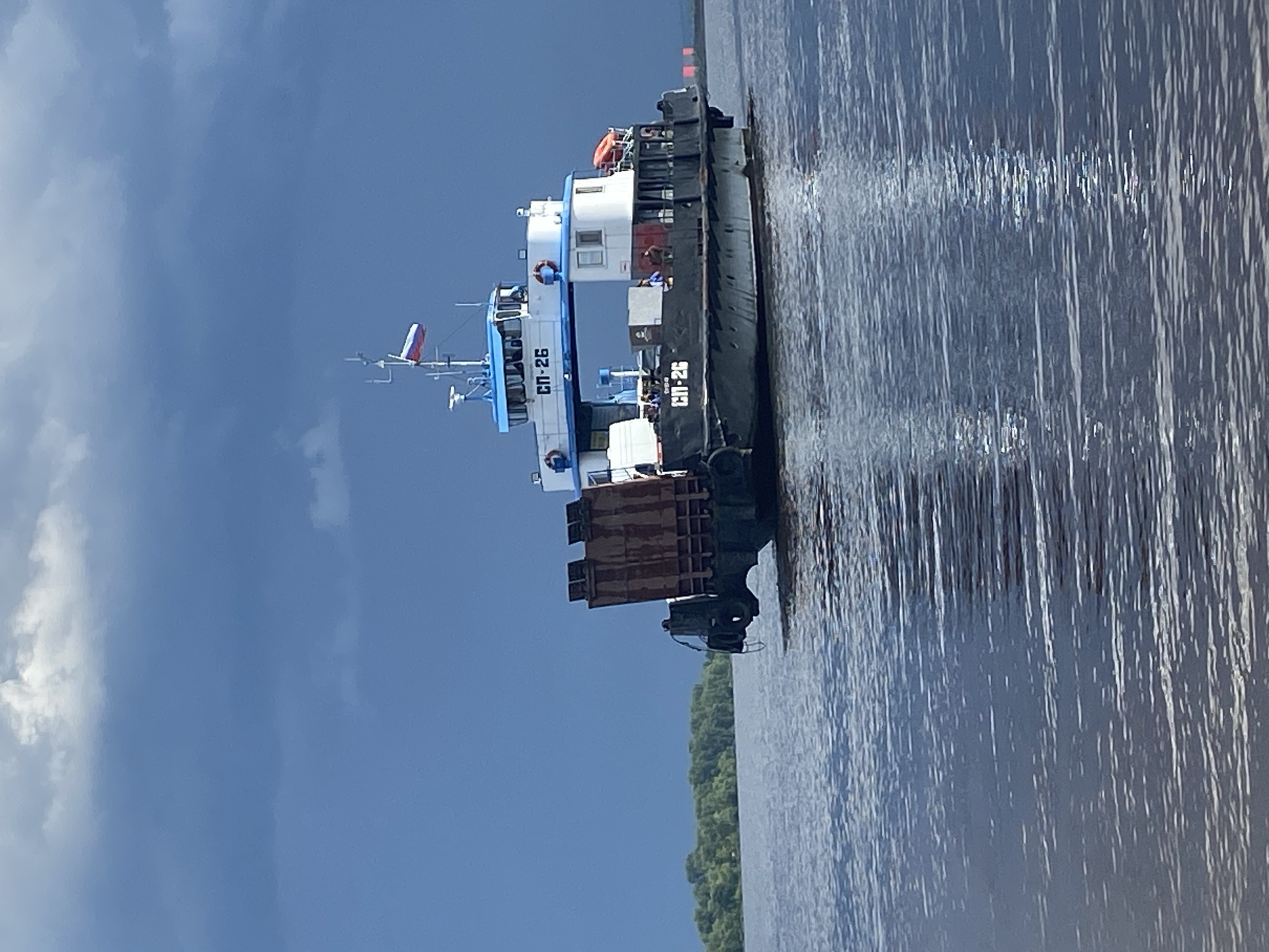 A barge is seen from the side, partially submerged in calm water. The barge is dark gray and has a white superstructure with blue accents. The text "СП-26" is visible in white on both the superstructure and the side of the barge. A small Russian flag is visible at the top of the superstructure. The barge appears to be carrying a load of what looks like industrial equipment, possibly scrap metal, on its deck. The sky is blue with scattered white clouds. The scene is likely set in the morning or early afternoon, given the lighting. The water is dark blue and reflects the sky and the barge. A thin line of green trees is visible along the horizon in the distance. The overall mood of the image is calm and serene.