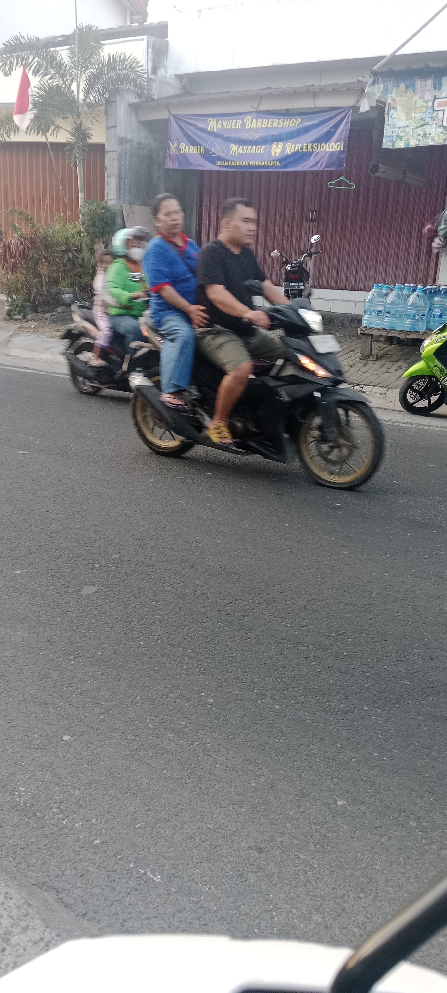 In Maguwoharjo, Indonesia, a busy street scene unfolds. Two motorcycles are the primary focus. The first, a black motorcycle, carries two individuals. A man in a black t-shirt and khaki shorts rides as the driver, while a woman in a blue shirt and jeans sits behind him. They are both in motion, suggesting they are traveling on the asphalt road. Another motorcycle, a vibrant green one, is parked on the right side of the frame. Behind the motorcycles, a building with a maroon-colored corrugated metal facade is visible. A blue banner hangs above the building's entrance, displaying text that reads "MANJER BARBERSHOP" in large letters, with smaller text below indicating "BARBER MASSAGE REFLEKSIOLOGI" and "JALAN PASEKAN YOGYAKARTA." To the left of the building, a palm tree with fronds and a red and white Indonesian flag are present. The environment appears to be an urban or semi-urban setting, with a road and buildings suggesting commercial activity. Several large blue water bottles are stacked on a wooden pallet in front of the building.
