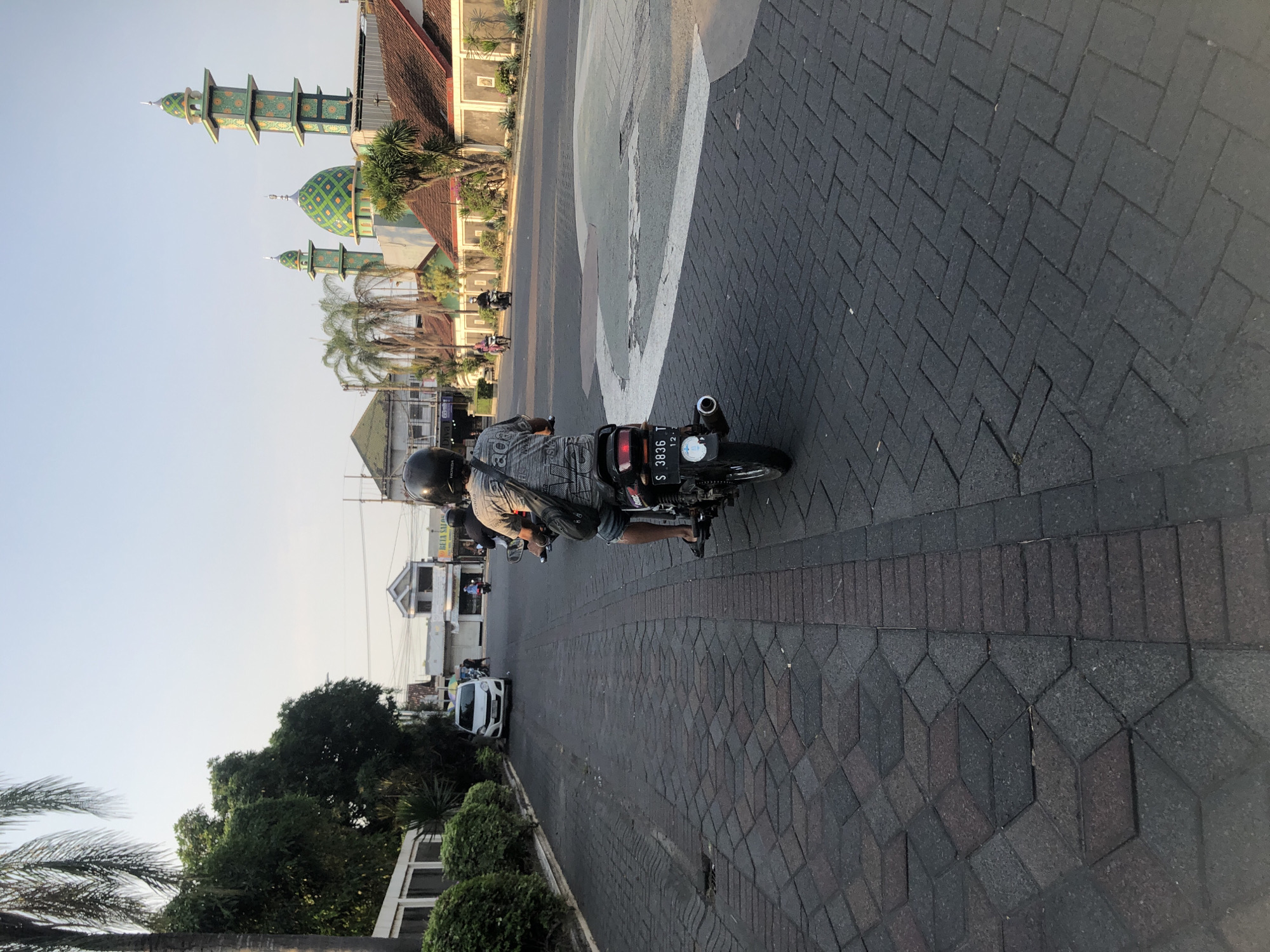 The image depicts a street scene in Boro, Indonesia, with a prominent mosque on the right side of the frame. The foreground shows a dark, patterned brick road leading towards the mosque and other buildings. A person is riding a motorcycle away from the viewer, wearing a helmet and a patterned shirt. Another motorcycle is visible further down the road. The mosque has a distinctive green dome and minarets, suggesting its architectural style. In the background, there are several buildings, some with red-tiled roofs, and lush green trees. The sky is clear and pale blue, indicating it is likely daytime. The overall impression is of a typical street in an Indonesian town, with religious and residential structures.