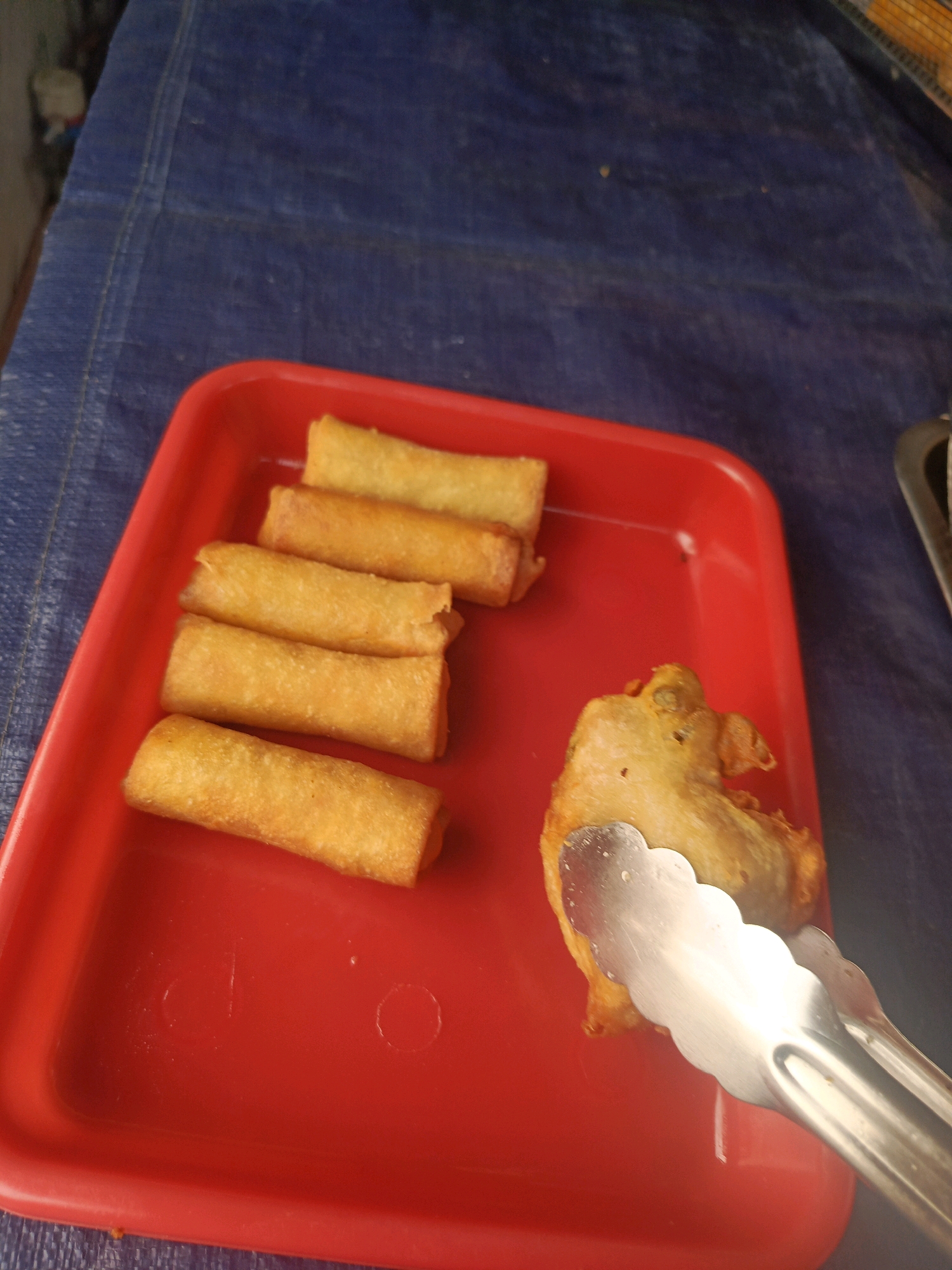 In Balaraja, Indonesia, a red plastic tray sits on a dark blue tarp. The tray contains several golden-brown spring rolls. To the right of the spring rolls, a piece of fried chicken is held by silver tongs. The background is a blurry dark blue tarp.