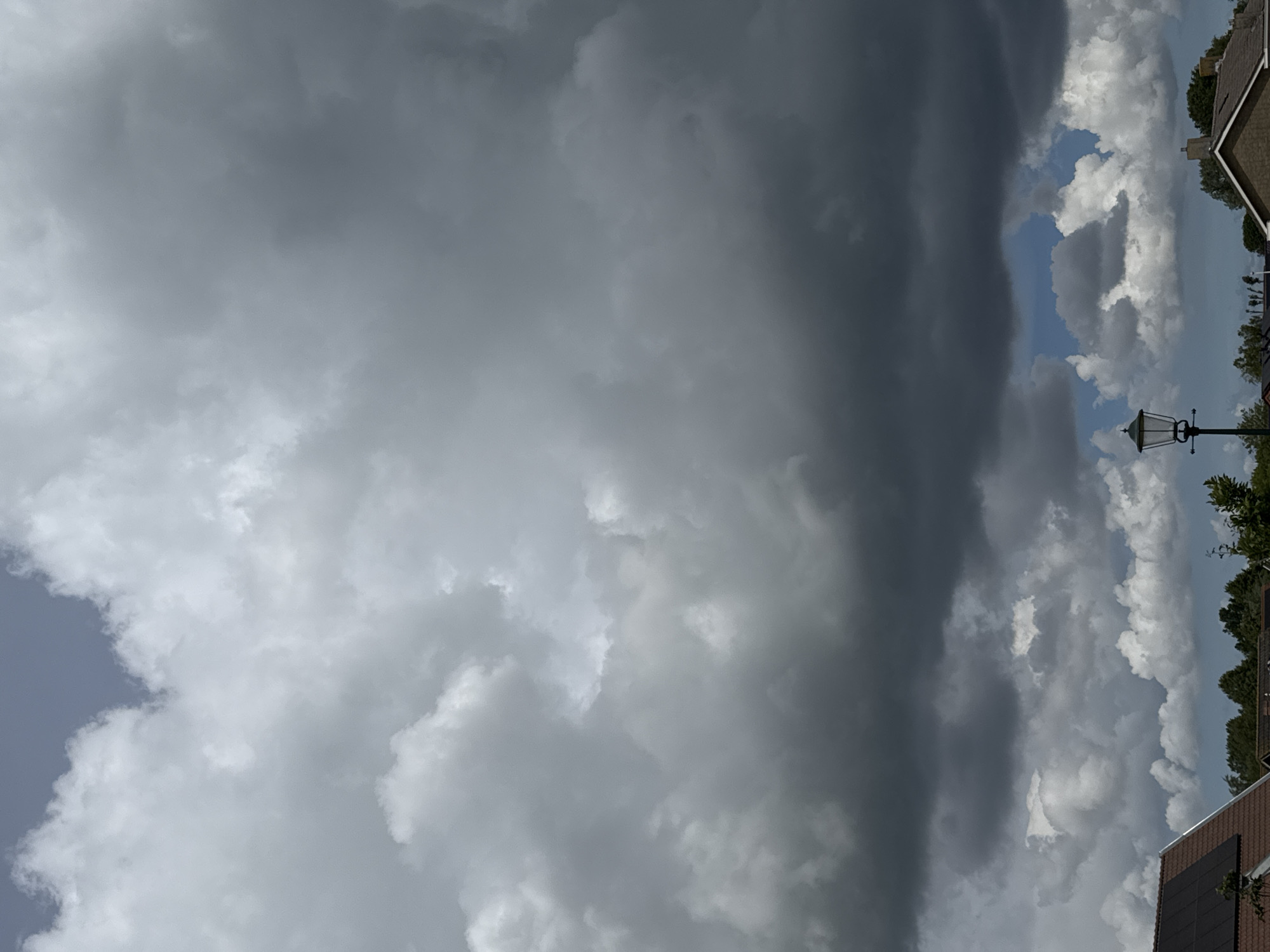 The image captures a cloudy sky above a residential area. The sky is dominated by large, dark grey clouds, with patches of lighter grey and white clouds interspersed. A small section of blue sky is visible in the distance, peeking through the clouds. Below the sky, the rooftops of houses and trees are visible. One house in the foreground has solar panels on its roof. A street lamp stands on the right side of the image. The scene appears to be outdoors, likely in a suburban or rural setting.