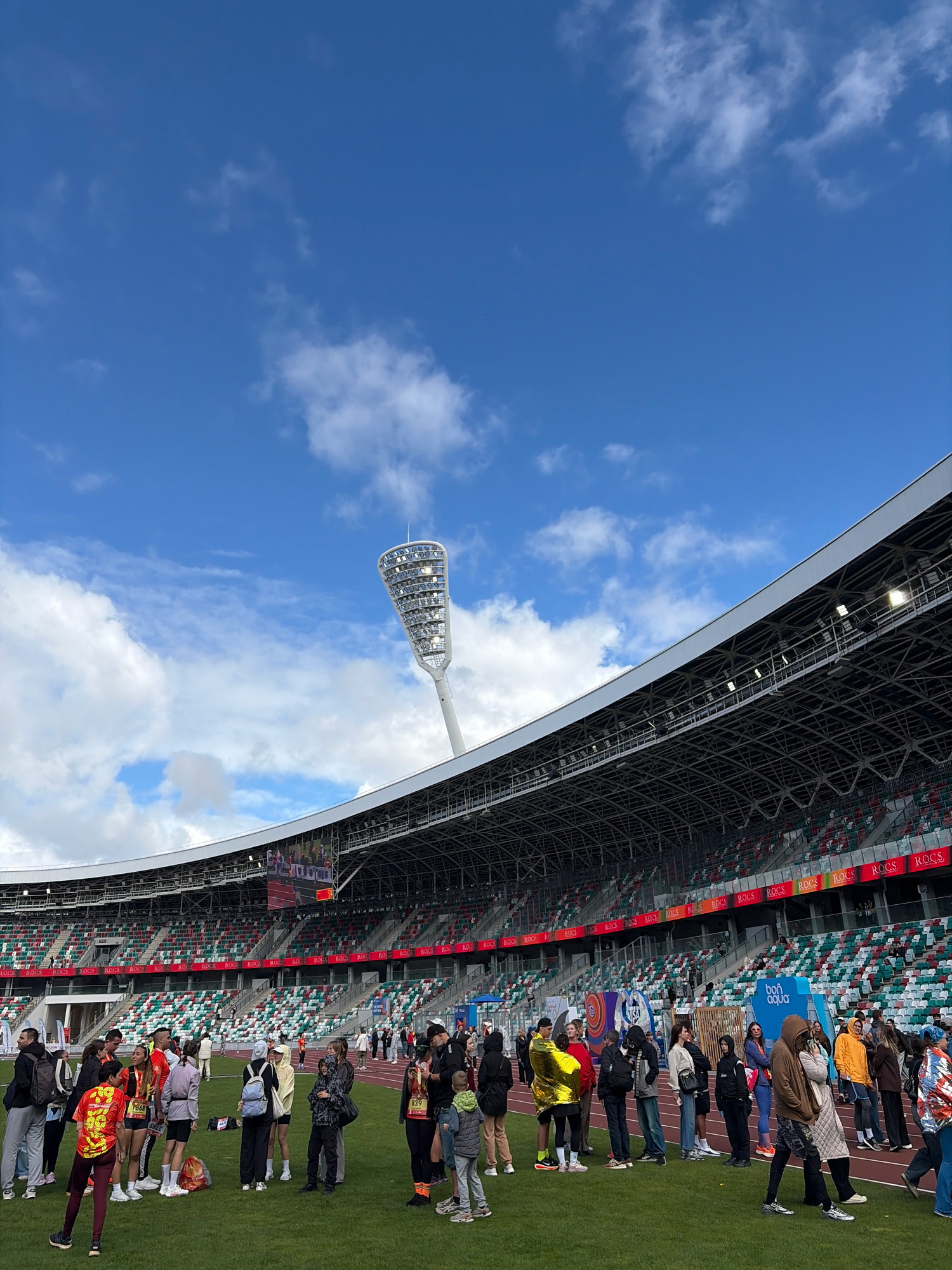 The image captures a scene at the Dinamo Stadium in Minsk, Belarus, under a bright blue sky with scattered clouds. A large crowd of people, many wearing athletic attire and some with medals, are gathered on the grassy field in front of the stadium's grandstands. The stadium itself features tiered seating in red, white, and green, with a prominent illuminated scoreboard visible in the background. A tall, modern lighting tower stands out against the sky to the right of the frame. Various banners and advertisements are displayed around the stadium, and people appear to be milling about, possibly before or after an event.