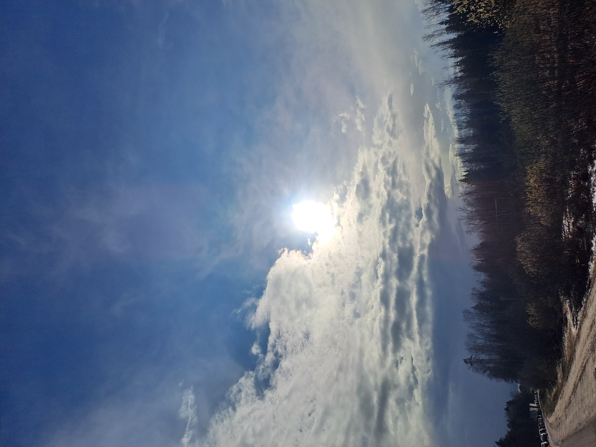 The image presents a daytime view in what is likely Babeevo, Russia, though there's no definitive way to confirm the city. The sky dominates the scene, showcasing a bright sun partially obscured by clouds, with varying shades of blue visible. The clouds appear to be a mix of cumulus and cirrus types.

On the right side of the frame, a line of trees suggests the edge of a forest or park area. A road or path can be seen on the lower right, near the trees, with several parked cars near it.