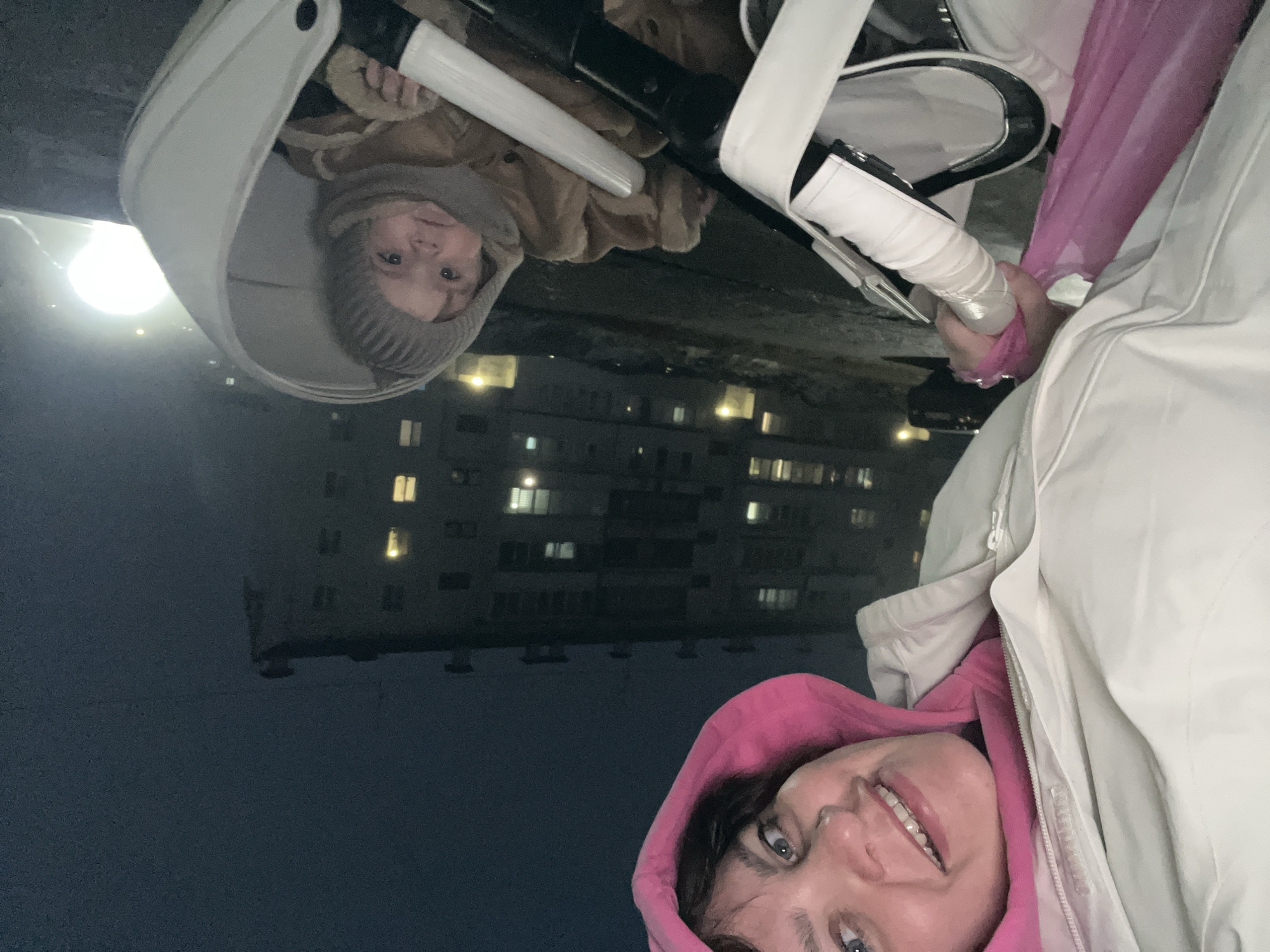A night-time selfie shows a woman in a pink hooded sweatshirt and beige jacket, standing in Noviy, Russia, with a baby in a stroller. The woman smiles at the camera. The baby, wearing a knitted hat and a brown jacket, looks up from the stroller. Streetlights are visible in the background, along with a block of apartments with lit windows.