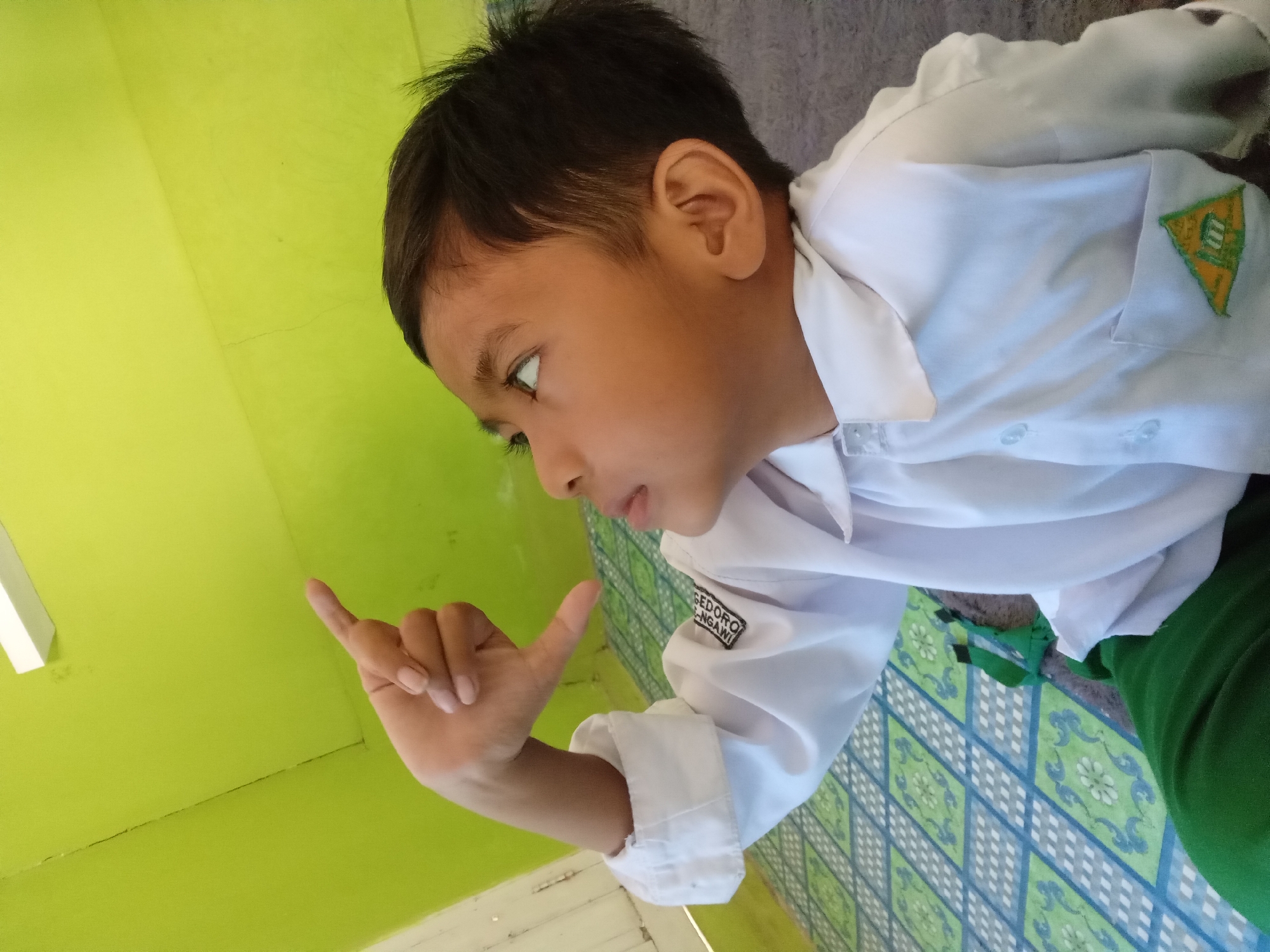 A boy in a white uniform shirt and green pants is sitting on a mat with a blue and green diamond pattern. His right hand is raised with his index finger and thumb forming an L-shape, and he is looking upwards. The background is a lime green wall. A white light fixture is visible at the top of the image. A patch with an emblem is visible on the left side of his shirt. The scene is likely indoors, given the wall and the presence of a mat. The image does not provide any specific clues about the location being Kedunggudel, Indonesia.