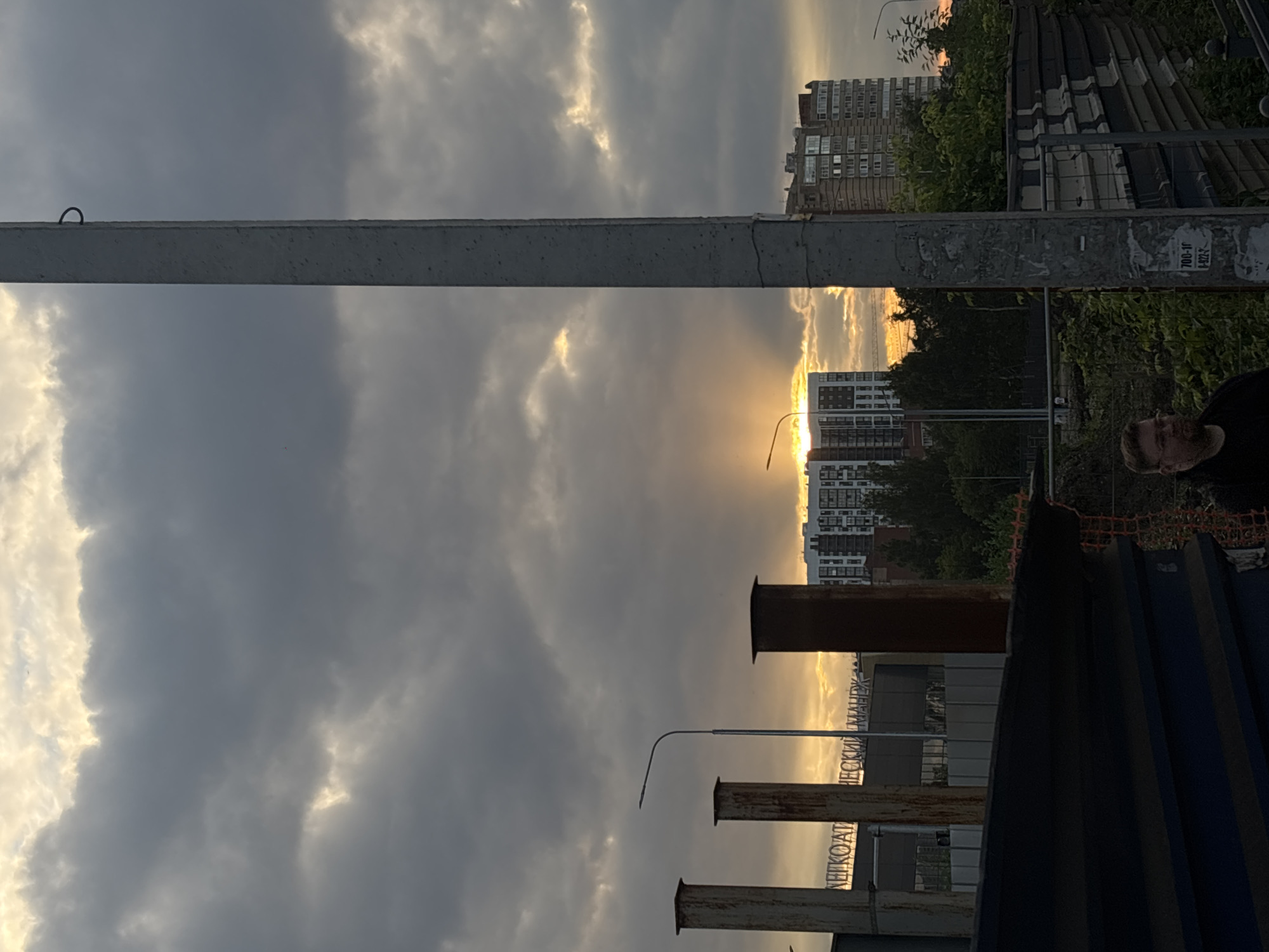The image depicts an urban sunset in Perm, Russia, with a sky filled with dramatic clouds illuminated by the setting sun. In the foreground, a large concrete pole stands prominently, obscuring part of the view. To the left, several industrial-looking structures with metallic or concrete surfaces rise vertically. Behind these, a modern building with numerous windows is visible, along with other residential buildings further in the background. Green trees and foliage are interspersed throughout the scene, suggesting a natural element within the urban landscape. A man wearing glasses and a dark top is partially visible in the lower right portion of the image. The overall impression is one of a transitional moment, with day giving way to evening in a developed area.
