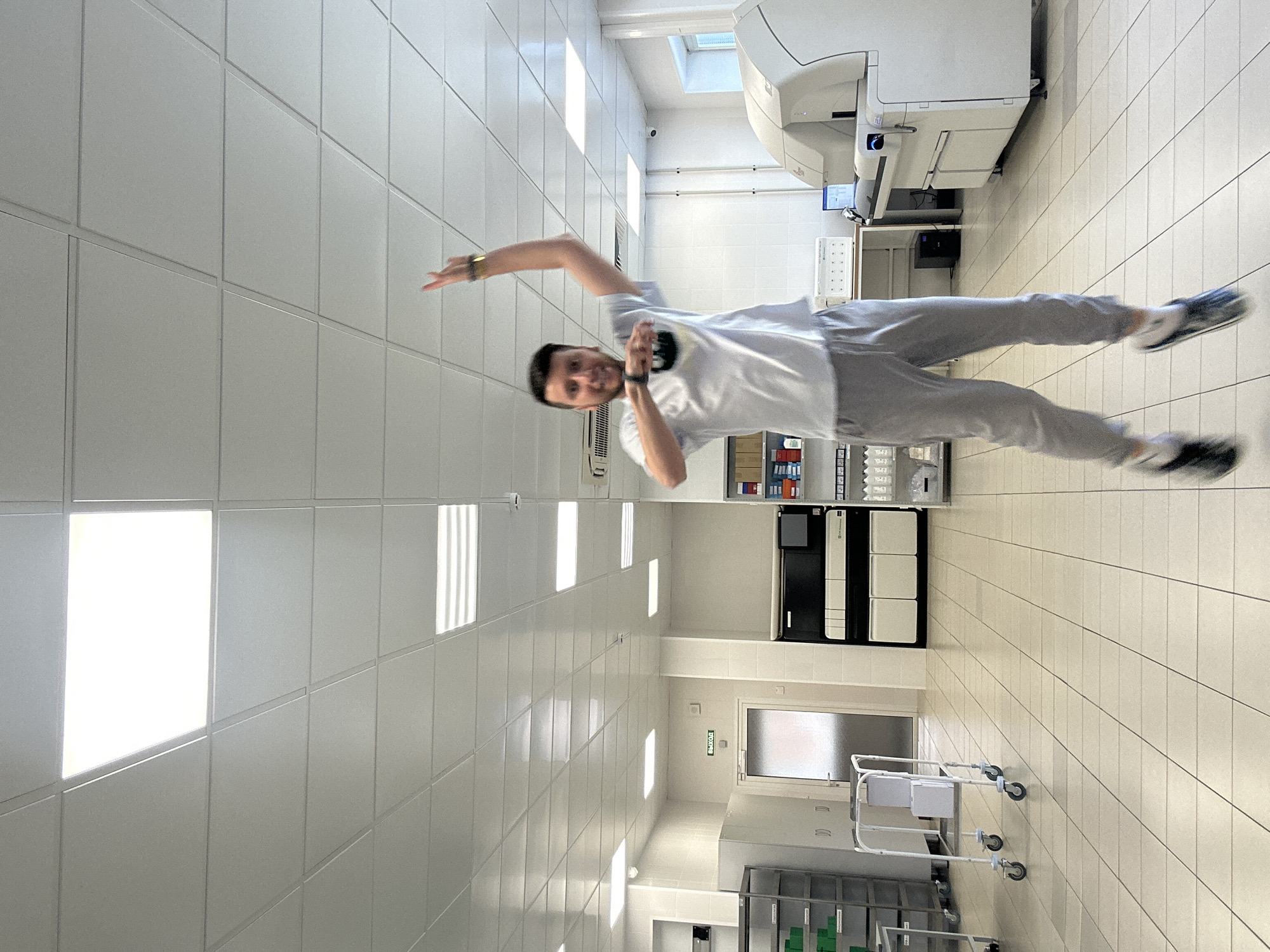 This image depicts the interior of a brightly lit laboratory or medical facility, identified by the "ВЫХОД" (EXIT) sign in Cyrillic, located in Moscow, Russia. A Caucasian male, appearing to be in his 20s or 30s with short dark hair and facial hair, is captured mid-jump, smiling broadly at the camera. He is wearing a light-colored t-shirt with a dark design on the chest pocket, light gray sweatpants, dark athletic shoes with white socks, a dark smartwatch on his left wrist, and a light bracelet on his right. His left arm is bent with his hand near his chest, while his right arm is raised above his head.
The environment features a grid-patterned light-colored tiled floor and a suspended ceiling with numerous bright, rectangular fluorescent or LED light panels that cast reflections. The walls are predominantly white, with sections of white square tiling visible.
To the left, a metal shelving unit holds green and white containers. A white medical cart with two white boxes rests nearby, adjacent to a white cabinet and a door with a frosted glass panel, above which the green "ВЫХОД" sign is mounted. An HVAC vent is visible on the wall in the upper-mid section. To the right, a large, multi-component white laboratory machine with dark accents is prominent, alongside a table holding a computer monitor. Further right, a shelving unit displays various laboratory supplies, and a security camera is mounted on the wall. A window is visible in the far right background.
