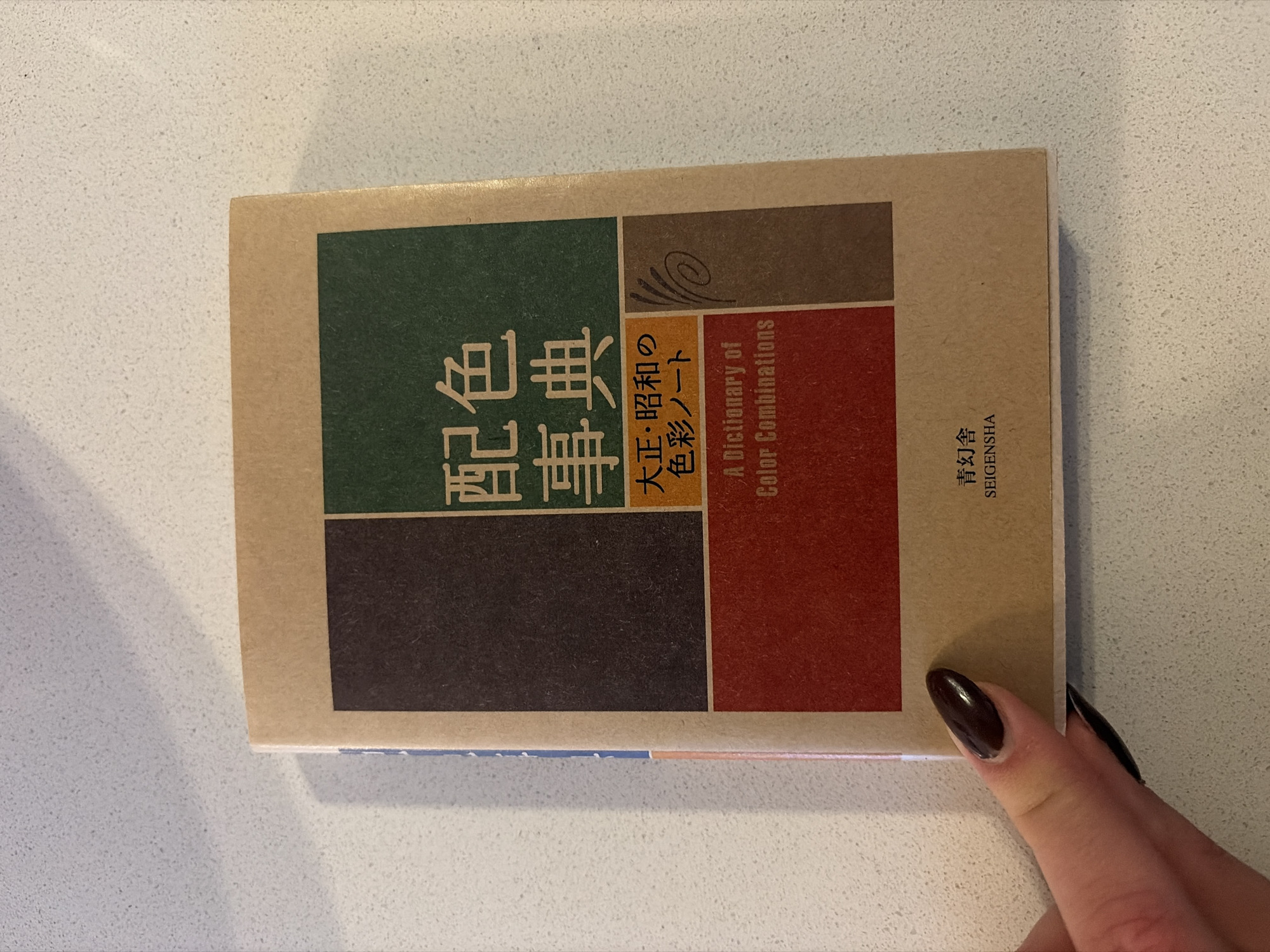 A close-up shot depicts a book titled "A Dictionary of Color Combinations." The book's cover is divided into rectangular blocks of green, brown, dark brown, and red against a beige background. Japanese text is prominently displayed, along with the English title. A hand with a dark, glossy nail is partially visible, touching the book. The book rests on a white countertop with subtle, speckled patterns. The photograph appears to be taken indoors with bright, diffused lighting. The book's publisher is listed as SEIGENSHA. There is no indication the image was taken in Montreal, Canada.