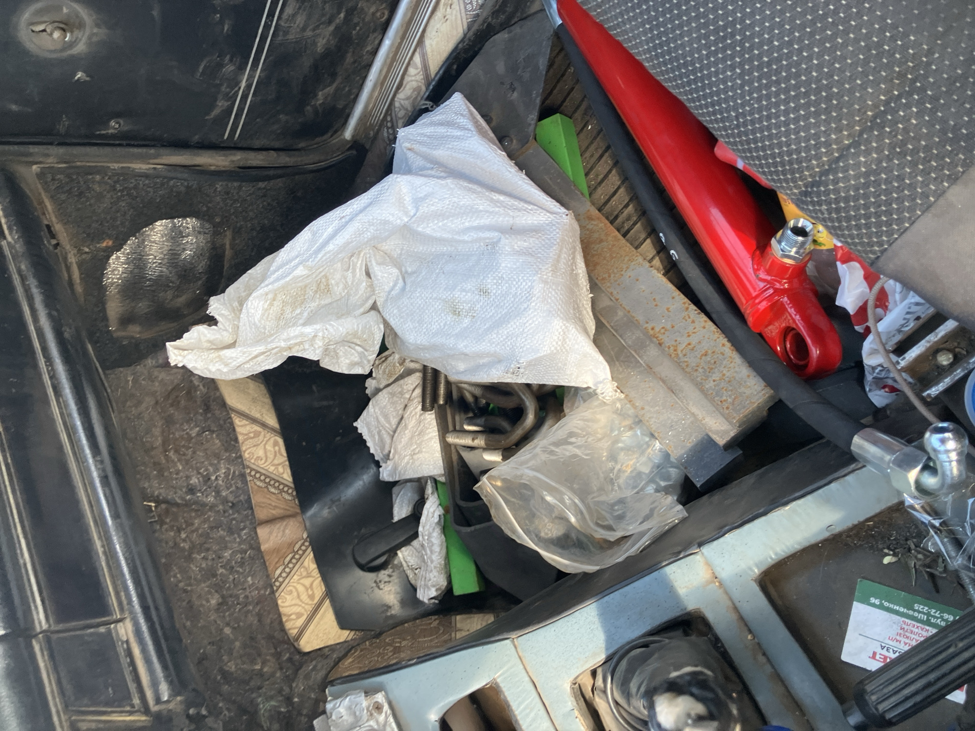 The image depicts the interior of a vehicle, likely a trunk or storage area, filled with various items. A large white plastic bag is prominent in the center, containing what appears to be crumpled paper or fabric. To the left, a blue plastic bin or compartment holds additional items, including a clear plastic bag with metal hardware, possibly U-bolts and nuts. A green object, possibly a tool or piece of material, is visible beneath the white bag and next to the blue bin. A red hydraulic cylinder is partially visible in the lower right quadrant of the image, with a black hose attached. Other metallic objects, including rusted metal, are scattered amongst the items. The environment appears somewhat cluttered and dirty, suggesting a workspace or storage area for tools and equipment. The location is identified as Molodizhne, Ukraine.