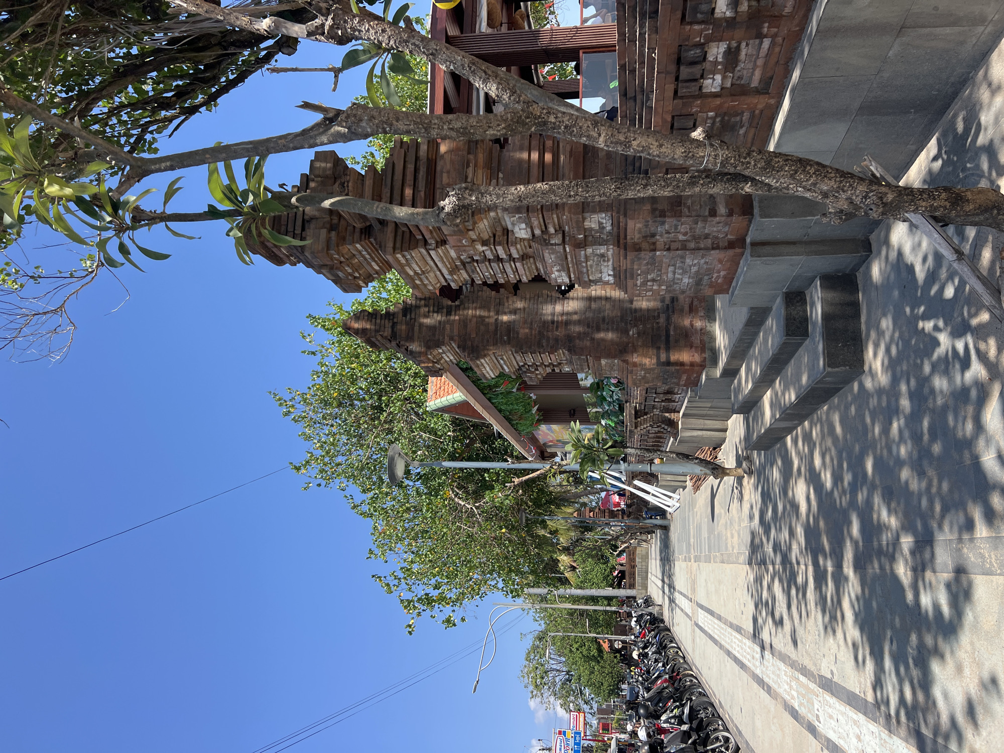 The image shows a sunny day in Kuta, Indonesia, with a clear blue sky. On the left side of the paved sidewalk, a row of parked motorcycles is visible, suggesting a busy commercial or tourist area. Further down the street, more trees and buildings line the road, indicating an urban environment.
On the right side of the sidewalk, a weathered brick structure, possibly an entrance or part of a building, stands out. A young tree with sparse leaves grows in front of the brickwork, its branches reaching towards the sky. The shadows of the trees are cast across the sidewalk, creating a dappled pattern.
In the background, more trees and greenery are seen, suggesting a tropical climate. The overall scene depicts a typical street in Kuta, a popular tourist destination known for its beaches and vibrant atmosphere. There are no people clearly visible in the foreground or mid-ground of the image, though a few figures may be present in the background, obscured by distance or foliage. The image is captured from a slightly low angle, looking down the sidewalk towards the left.