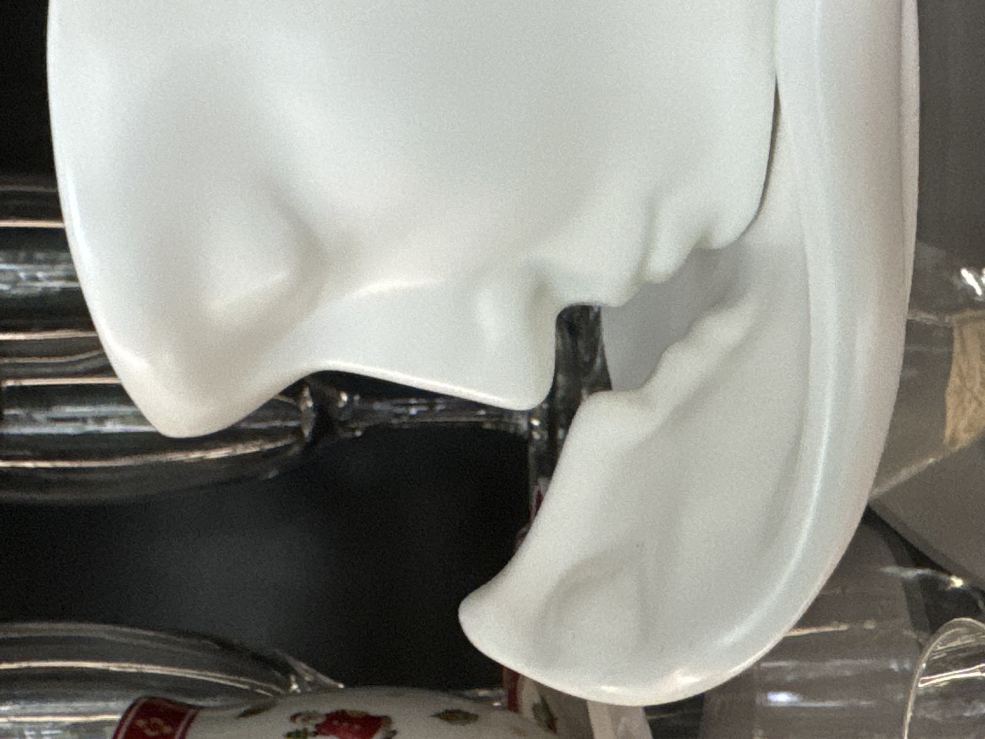 The image shows a close-up of a white ceramic sculpture of a face, with its nose and lips pressed against a smooth, curved surface. The sculpture is on a saucer or base, also white. In the background, out of focus, are clear glass objects, possibly wine glasses, and a dark, textured surface. A small portion of a white and red decorative object is visible on the far left. The lighting is soft, creating subtle shadows on the sculpture. The location in Steklyannyi, Russia is not discernable from the visual information provided.
