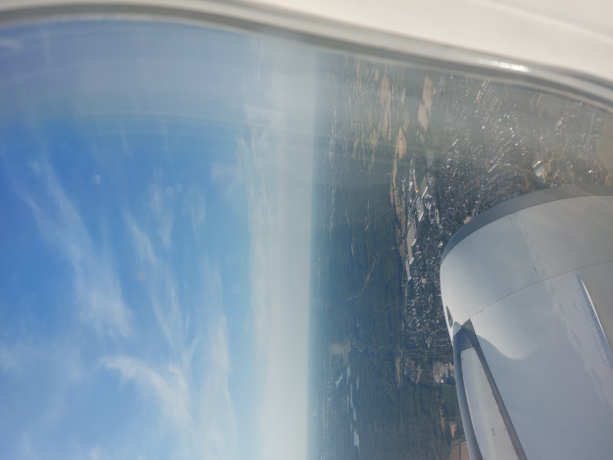 This is an aerial photograph taken from an airplane window, looking out over the city of Ratingen, Germany. The foreground is dominated by the engine nacelle of the aircraft. Beyond that, the landscape unfolds with a mix of dense green forests, bodies of water, and a sprawling urban area. The city appears to be a suburban landscape with houses, roads, and industrial buildings visible. The sky above is a clear blue with scattered wispy clouds. The lighting suggests it is daytime, likely mid-morning or afternoon, given the shadows cast on the land. There are no people or animals visible in the image. The overall impression is of a landscape viewed from a high altitude.