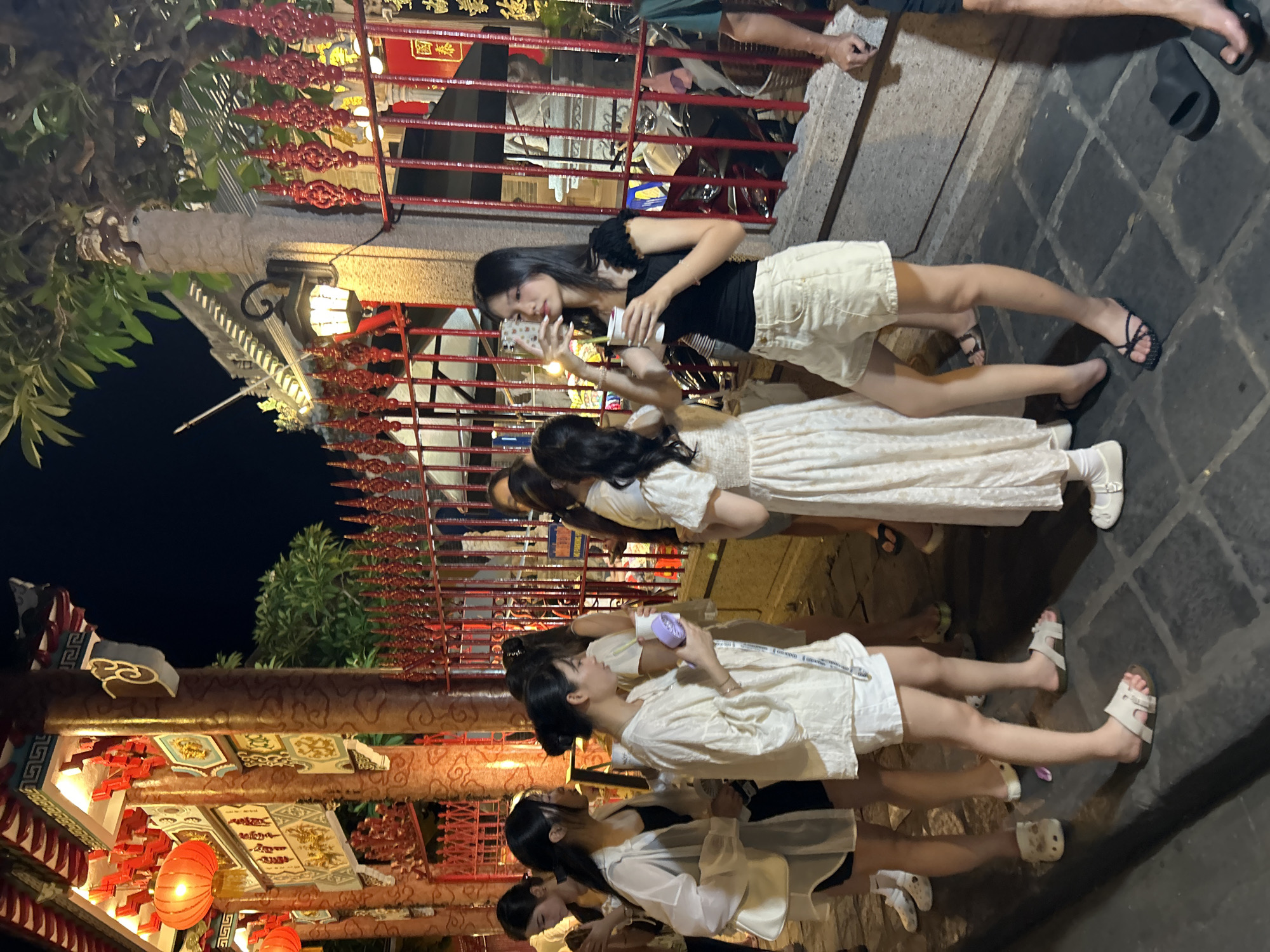 In Hội An Ward, Vietnam, a group of young women are gathered at night in front of a vibrantly decorated building, possibly a temple or a traditional establishment, characterized by red ornate fences and glowing lanterns. The architectural style suggests a strong East Asian influence. The women, dressed in light-colored casual summer clothing, are interacting with each other. One woman in the foreground is holding a cup and a phone, appearing to take a selfie. Another woman next to her is holding a purple cup. The ground is paved with stone tiles. The scene is illuminated by artificial lights, creating a warm and lively atmosphere.