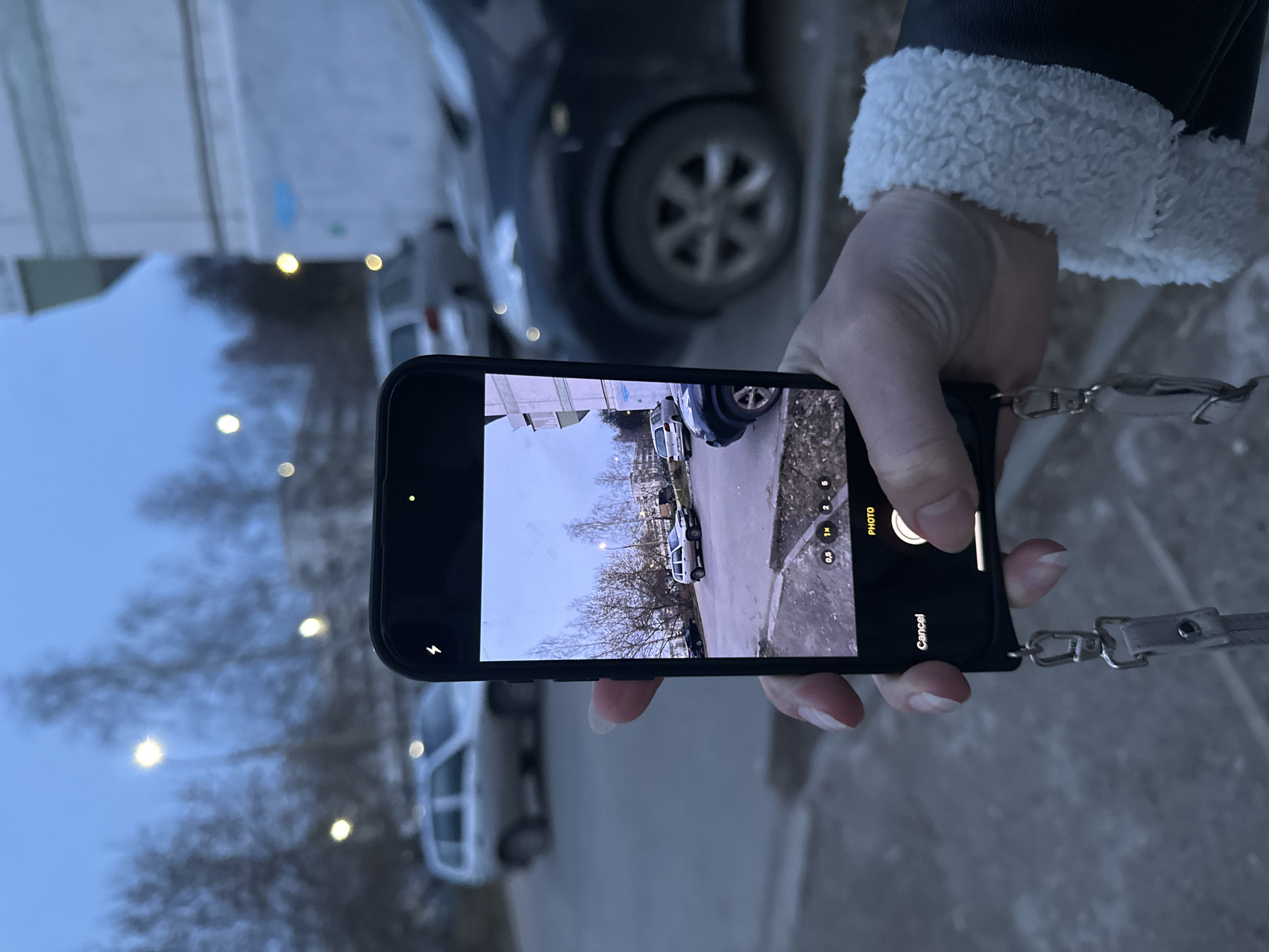 The image captures a person's hand holding a black smartphone, seemingly taking a photo in an outdoor setting in Chaikovsky, Russia. The person is wearing a dark jacket with a white, fluffy sherpa-style cuff, and their fingernails are painted a light, possibly nude or pale pink color. The phone is encased in a black protective cover and features a light-colored lanyard or strap attached to its bottom right corner.
The smartphone's screen displays its camera interface, showing a live preview of the scene the person is attempting to capture. This preview depicts a street with at least two parked cars – one white van/minivan and another darker vehicle – alongside bare trees and an overcast sky. The camera mode is set to "PHOTO," with zoom options "0.5", "1X", "2", and "5" visible on the right side of the screen. A flash icon is present on the left, and a "Cancel" button is near the bottom right. A green dot is visible in the top left corner of the phone screen, typically indicating active camera usage.
The background of the overall image, beyond the person and the phone, is out of focus. It shows blurred elements consistent with an urban or suburban environment, including parts of buildings, other indistinct vehicles, and scattered glowing lights that might be streetlamps or interior lights, suggesting a gloomy day, dusk, or early evening. The overall lighting is soft and diffused, indicating an overcast sky.