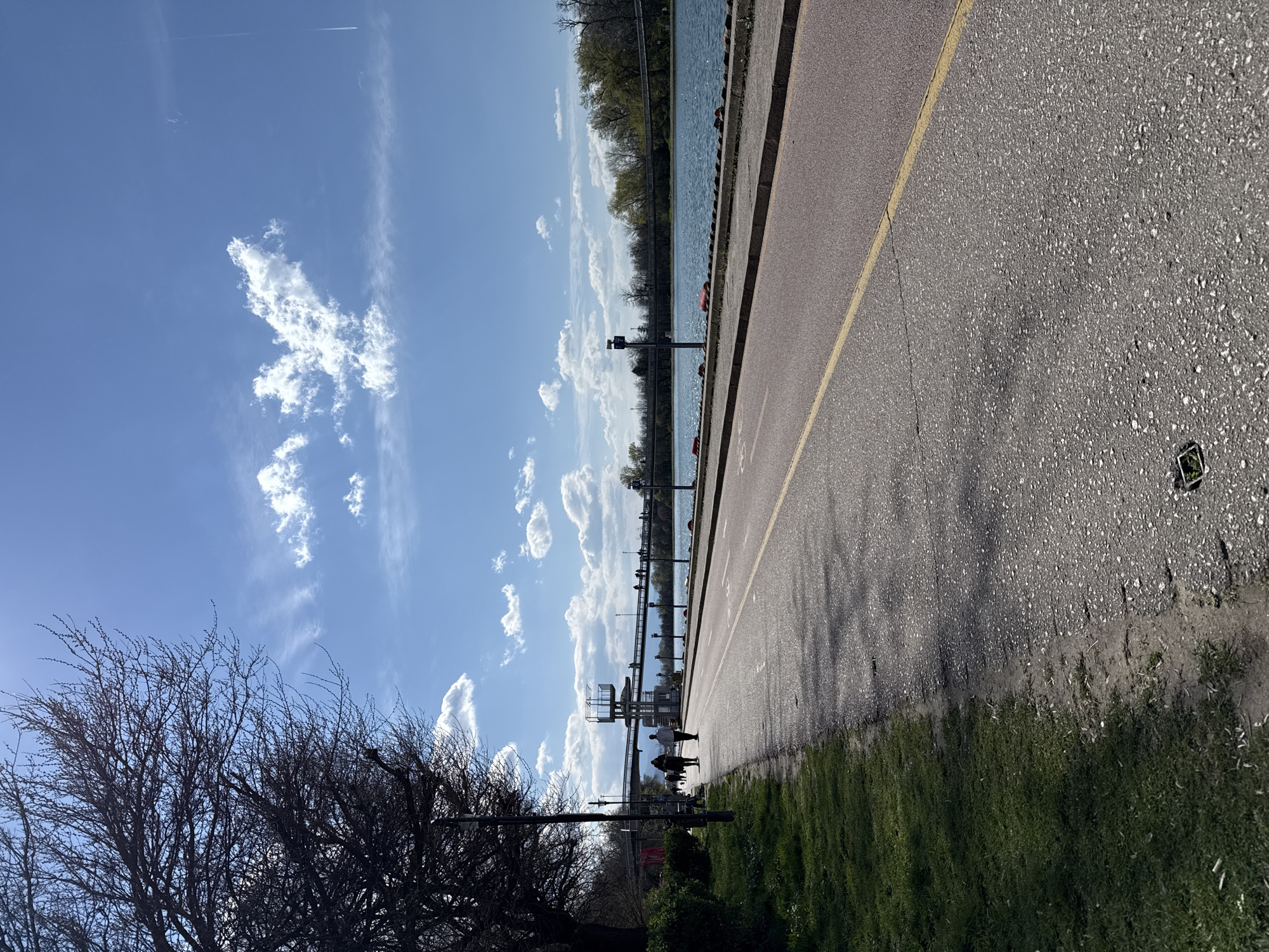The image shows a horizontal view of a paved area near a body of water under a bright blue sky with scattered cumulus clouds. A yellow line demarcates the edge of the pavement. To the left of the pavement is a grassy area with a leafless tree visible in the foreground.
In the distance, along the water's edge, are poles or structures, some resembling light posts or support towers, set against the water and treeline on the opposite shore. Several people are walking along the edge of the pavement near the structures.
Based on the location characteristics, particularly the presence of the water body, tree types, and structure placement, the image is likely taken near the Rowing Channel (Grebnia Kanal) in Plovdiv, Bulgaria. The Rowing Channel is a popular recreational area that includes a paved path alongside the water, making it consistent with the image.