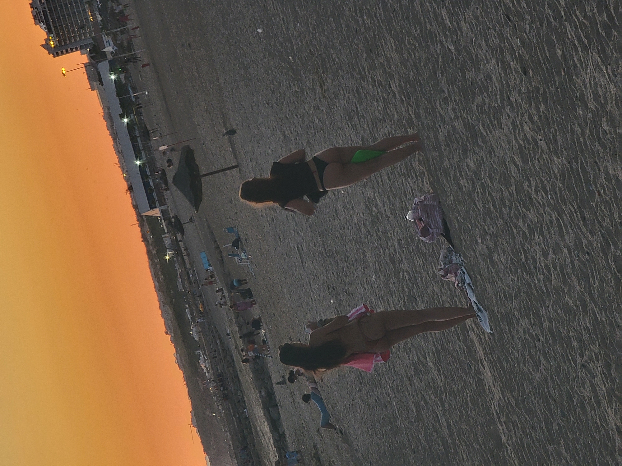 The image depicts a sandy beach scene at Quarteira, Portugal, during sunset. The sky is a vibrant orange. In the foreground, two women are visible from behind, walking on the sand. The woman on the left is wearing a bikini and a pink garment draped over her arm, while the woman on the right is wearing a black top and a bikini bottom, holding a green object. Several beach umbrellas, lounge chairs, and other beachgoers are scattered across the beach in the midground and background. Buildings and structures, likely hotels and apartments, are visible in the distance, with some illuminated lights.