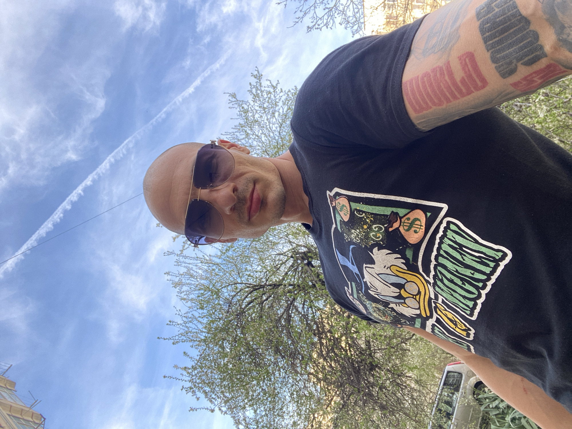 The image presents a bald-headed man wearing sunglasses and a black t-shirt with a cartoon graphic. He stands outdoors on what appears to be a sunny day, with blue skies and contrails visible above. The background includes foliage from trees. Part of a white vehicle is visible to the lower right. Based on visual assessment, the location appears to be Pleven, Bulgaria. This cannot be confirmed definitively without additional information. The subject is looking directly at the camera. His left arm, visible in the upper right corner, has extensive tattoos.