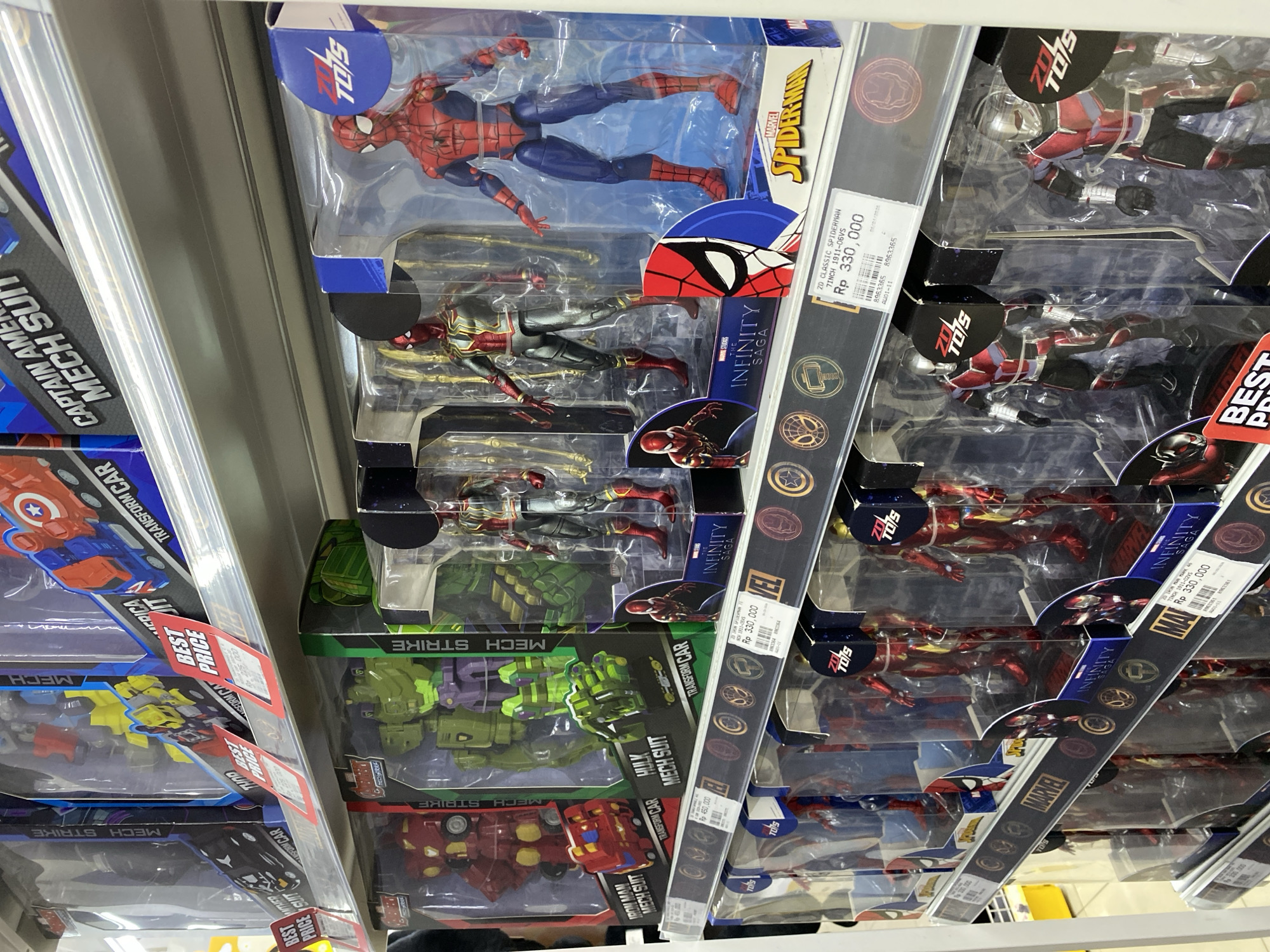 The image shows a retail store shelf displaying action figures of Marvel characters in Banjarmasin, Indonesia. The figures, including Spider-Man, Iron Man, and the Hulk, are packaged in clear plastic boxes with accompanying branding and pricing information. The shelves are well-lit, and the products are neatly arranged, suggesting a typical retail environment. The background shows more shelves stocked with similar merchandise, indicating a large selection of toys available. The overall scene is one of commercial activity, with a focus on popular superhero collectibles.