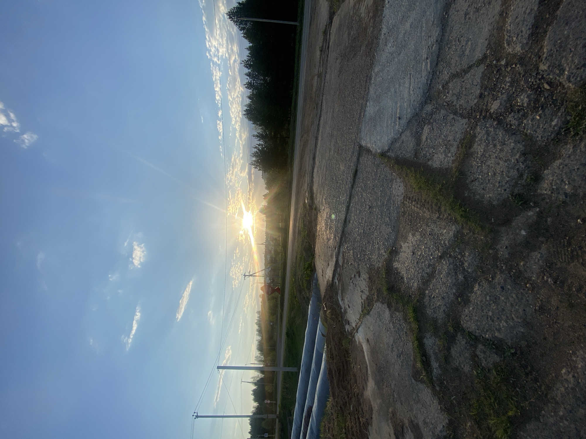 The image captures a rural roadside scene at sunrise or sunset, likely in a temperate climate. The sky transitions from a clear blue to a brighter, sunlit horizon with scattered clouds. The sun's rays shine intensely. Power lines and utility poles are visible extending along the road. On the right side of the image, a weathered concrete surface with some grass growing through the cracks dominates. Adjacent to the concrete, large pipes run parallel to the road. Beyond the utility lines and pipes is a field of grass, and the edge of a dense treeline marks the horizon. There are no people visible in the scene.