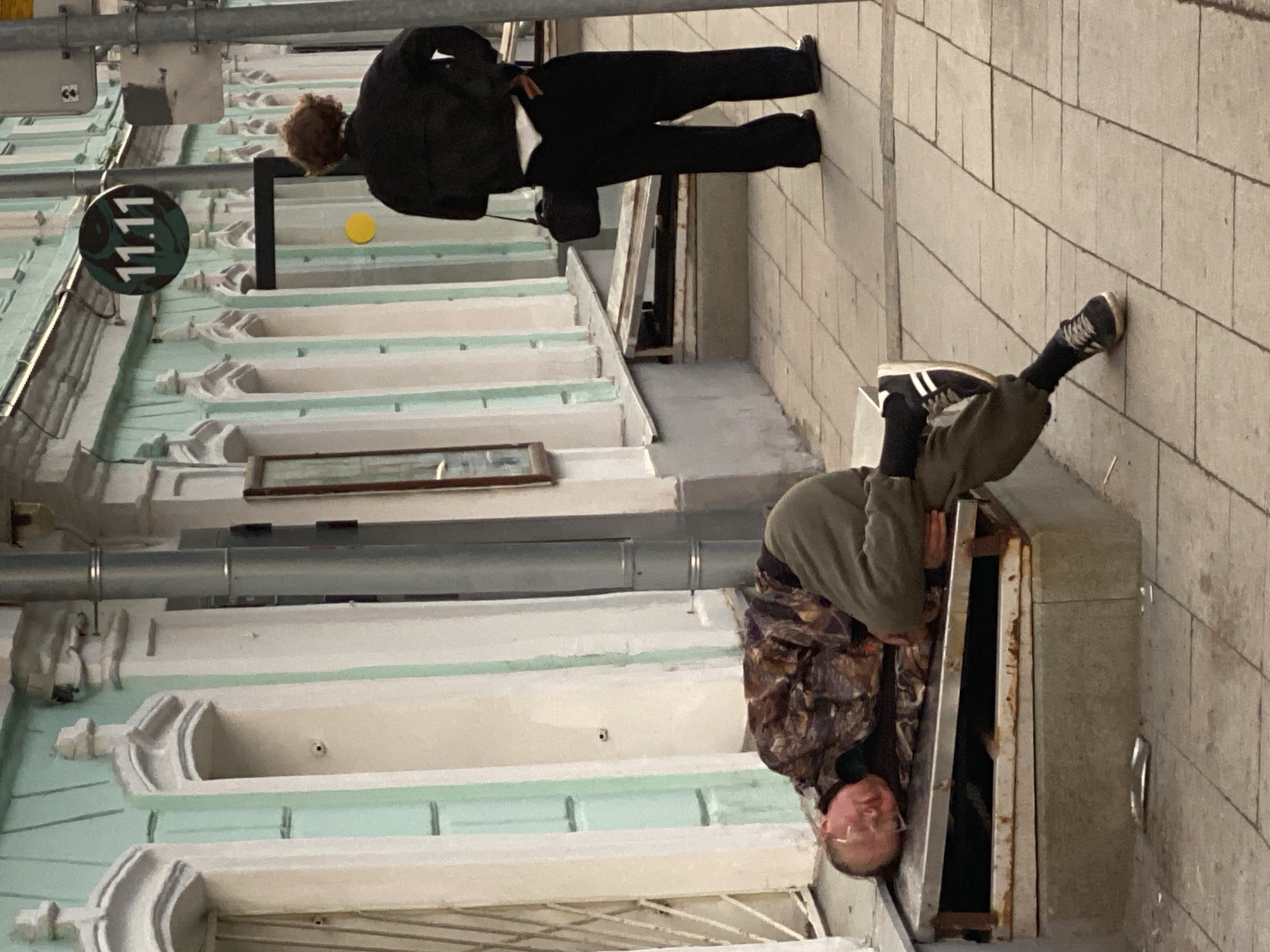 The image depicts a scene in Moscow, Russia. An older man, wearing glasses and a camouflage jacket, is partially inside a utility access point on the sidewalk. A person in a dark suit stands on the sidewalk near a building with ornate window ledges. A sign that reads "11.11" is visible.