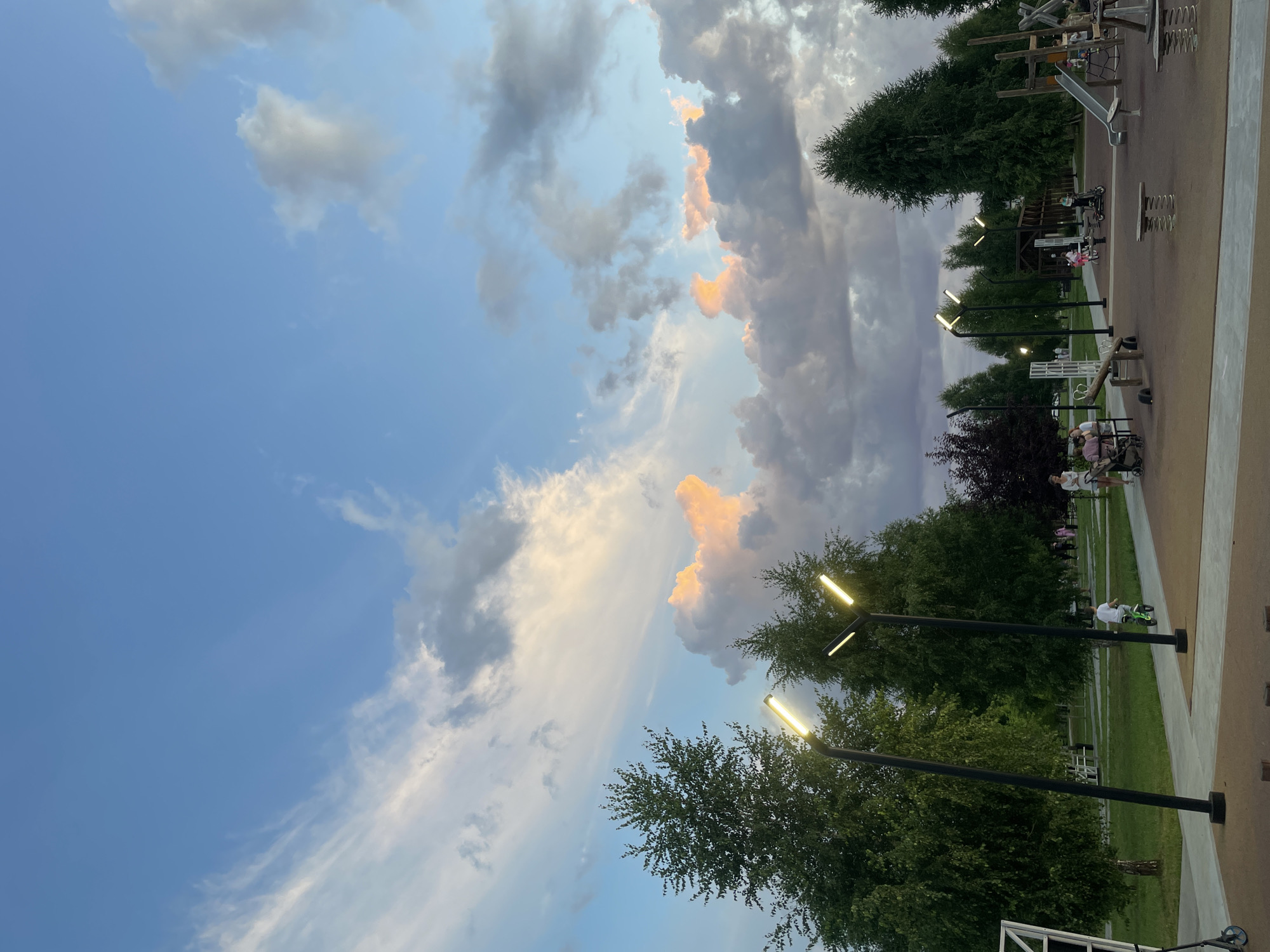 The image depicts a playground in the city of Laishevo, Russia, during the evening. The sky is a mix of blue and colorful clouds, with some clouds tinged pink and orange from the setting sun. Tall, slender trees line the perimeter of the playground, and modern streetlights with yellow bulbs illuminate the scene. The playground itself is equipped with various structures, including slides, swings, and climbing frames. Several children are visible, some riding bicycles and scooters, while others are playing with their parents. The ground of the playground is covered with a soft, brown material. The overall atmosphere appears peaceful and family-friendly, suggesting a pleasant evening spent outdoors.