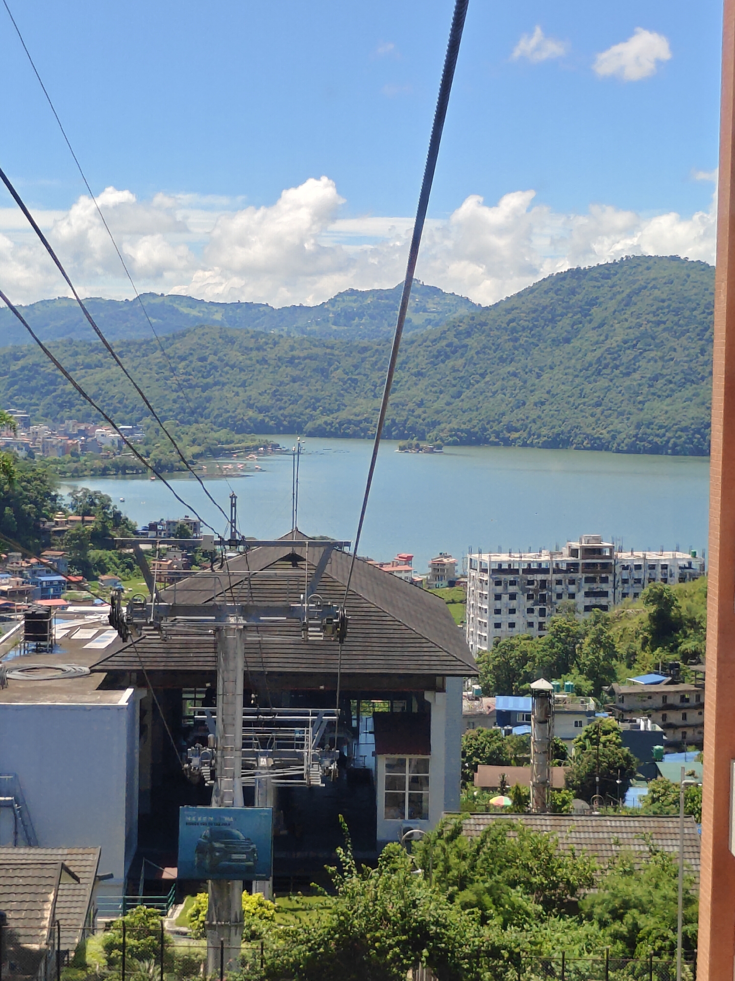 This image captures a view of the Phewa Lake and surrounding hills in Pokhara, Nepal, likely from a cable car station. In the foreground, a structure with a dark, sloped roof and metal supports is visible, part of a cable car system. A sign advertising a car is affixed to one of the support poles. Beyond this, Phewa Lake stretches out, a body of calm, blue water dotted with several small boats. The far side of the lake is lined with buildings, indicating a town or city. Lush green, tree-covered hills form the backdrop, rising to mountains in the distance under a bright blue sky with scattered white clouds. The overall scene depicts a scenic, urban and natural landscape.