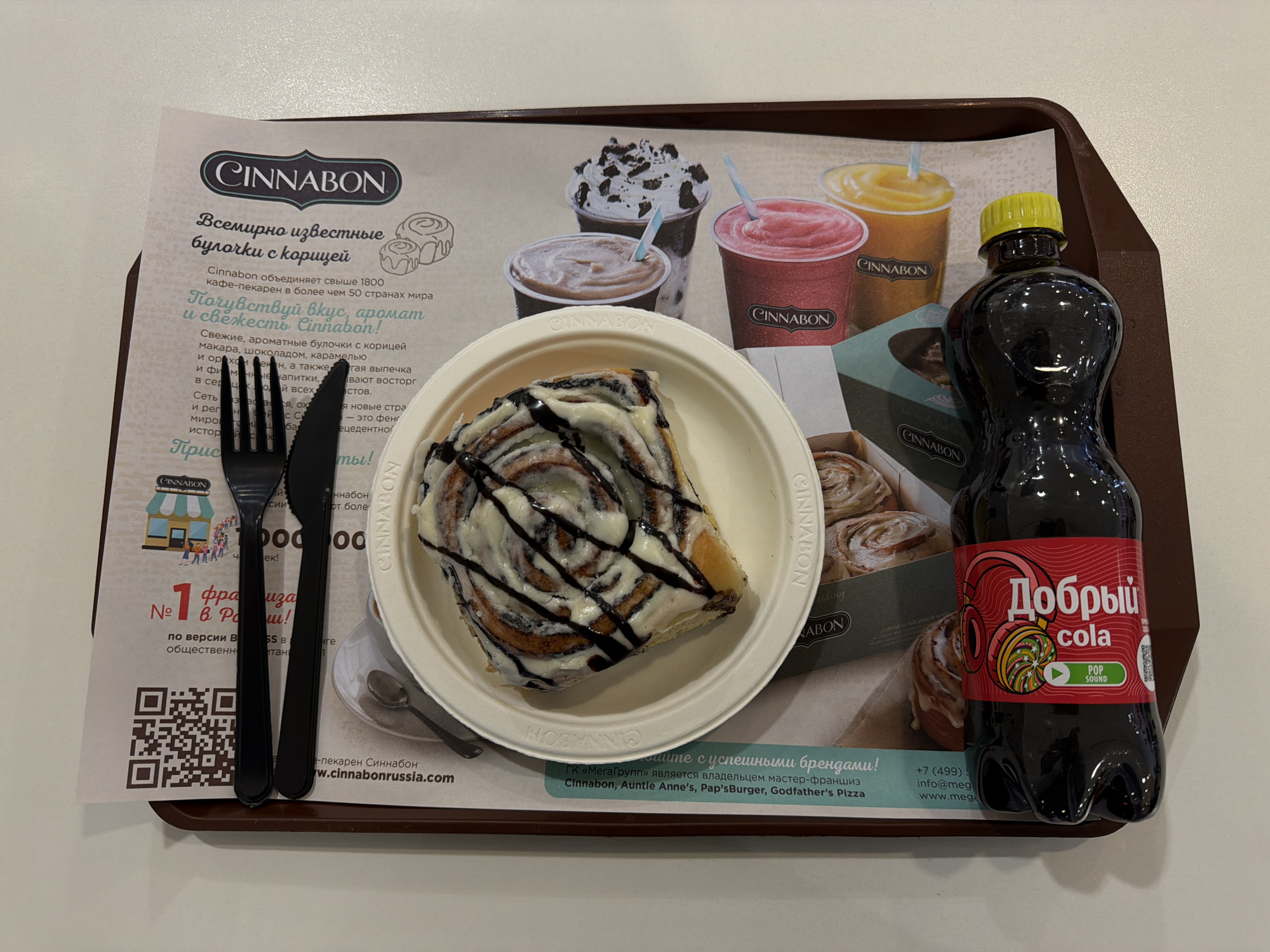 The image depicts a tray holding a Cinnabon cinnamon roll, a bottle of "Добрый Cola" (a Coca-Cola substitute produced in Russia), and a knife and fork. The cinnamon roll is in a white plate with white and dark chocolate topping, while the Cola bottle is unopened and has a yellow cap and a red label. The entire arrangement sits on a paper placemat featuring Cinnabon branding, images of other food and drink items, and a QR code. The placemat also includes text in Russian about the Cinnabon brand. The setting appears to be a Cinnabon cafe, as indicated by the placemat and the availability of a cinnamon roll and a cola-like beverage. It is implied this is in Krasnoyarsk, Russia given the Cinnabon branding on the placemat and the "Добрый Cola" branding.