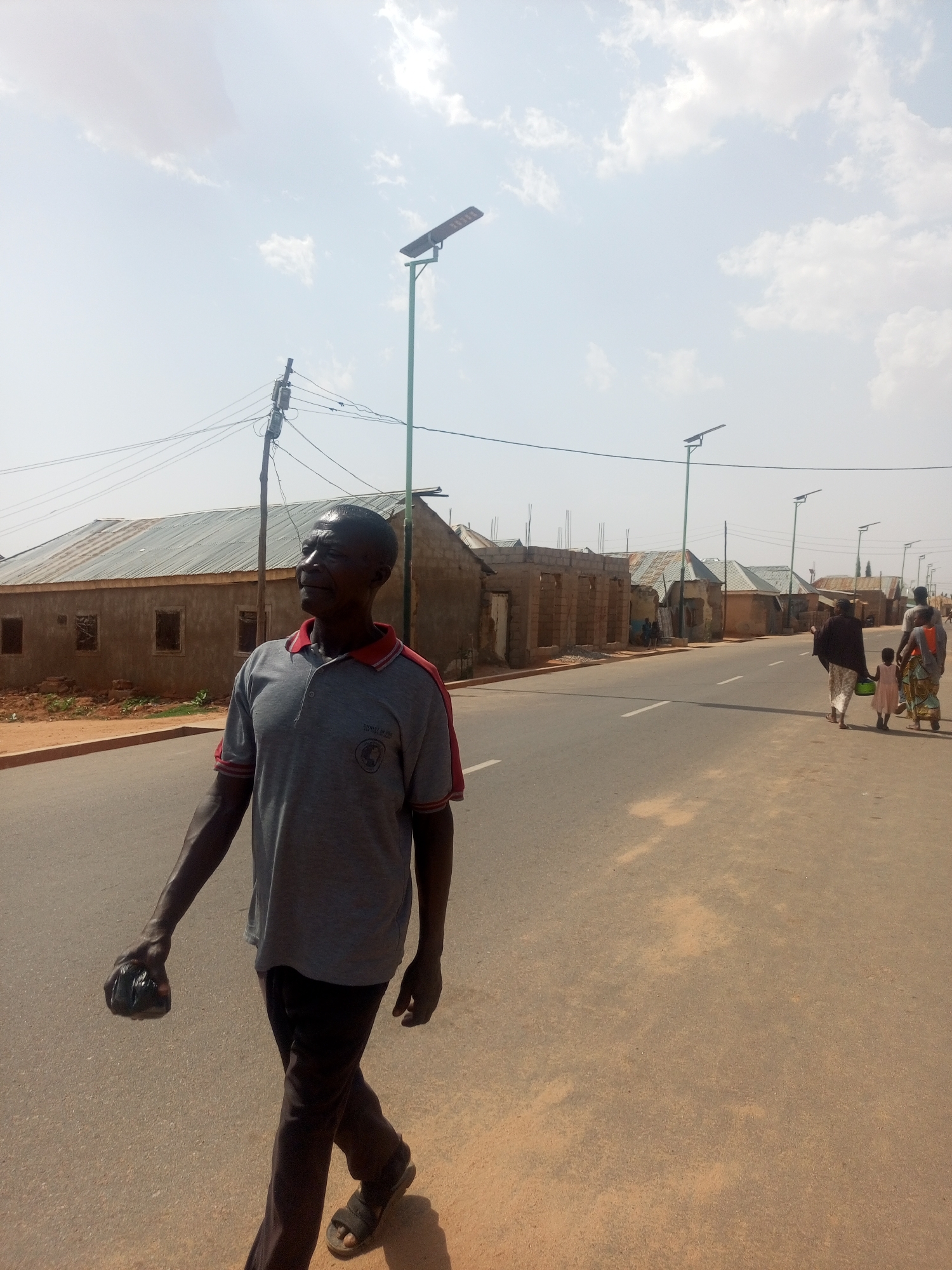 The image shows a street in Gombe, Nigeria. A middle-aged man walks on the right side of the road, wearing a grey shirt with red trim, dark pants, and sandals. He is carrying a black object in his left hand. In the background, a group of women and children walk on the right side of the road, away from the camera. The street is paved and lined with low, single-story buildings, some of which appear to be under construction or in disrepair. Utility poles and solar streetlights are visible along the road. The sky is partly cloudy.