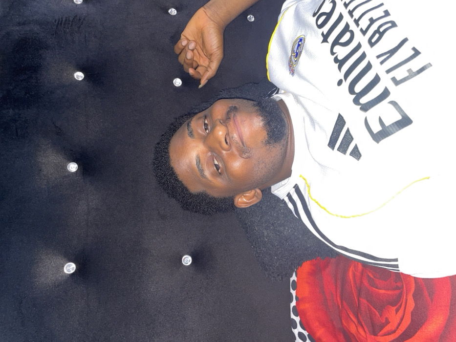 This image depicts an adult male indoors in Ube, Nigeria. The male, with dark skin, short, dark, curly hair, and a goatee-style beard, is positioned against a dark, possibly velvet, tufted headboard or wall panel adorned with clear, faceted buttons arranged in a grid pattern. He is wearing a white short-sleeved sports jersey featuring black Adidas stripes on the shoulders and a Real Madrid crest on the left chest. The jersey prominently displays the sponsor text "Emirates FLY BETTER" in black lettering. His left hand is partially visible in the upper right quadrant of the frame, with fingers resting against the dark background. A portion of a patterned pillow or blanket, displaying a large red rose design on a red background, and a small section of black and white polka dots, is visible in the lower-left corner of the frame. The male exhibits a slight smile and is looking directly towards the camera.