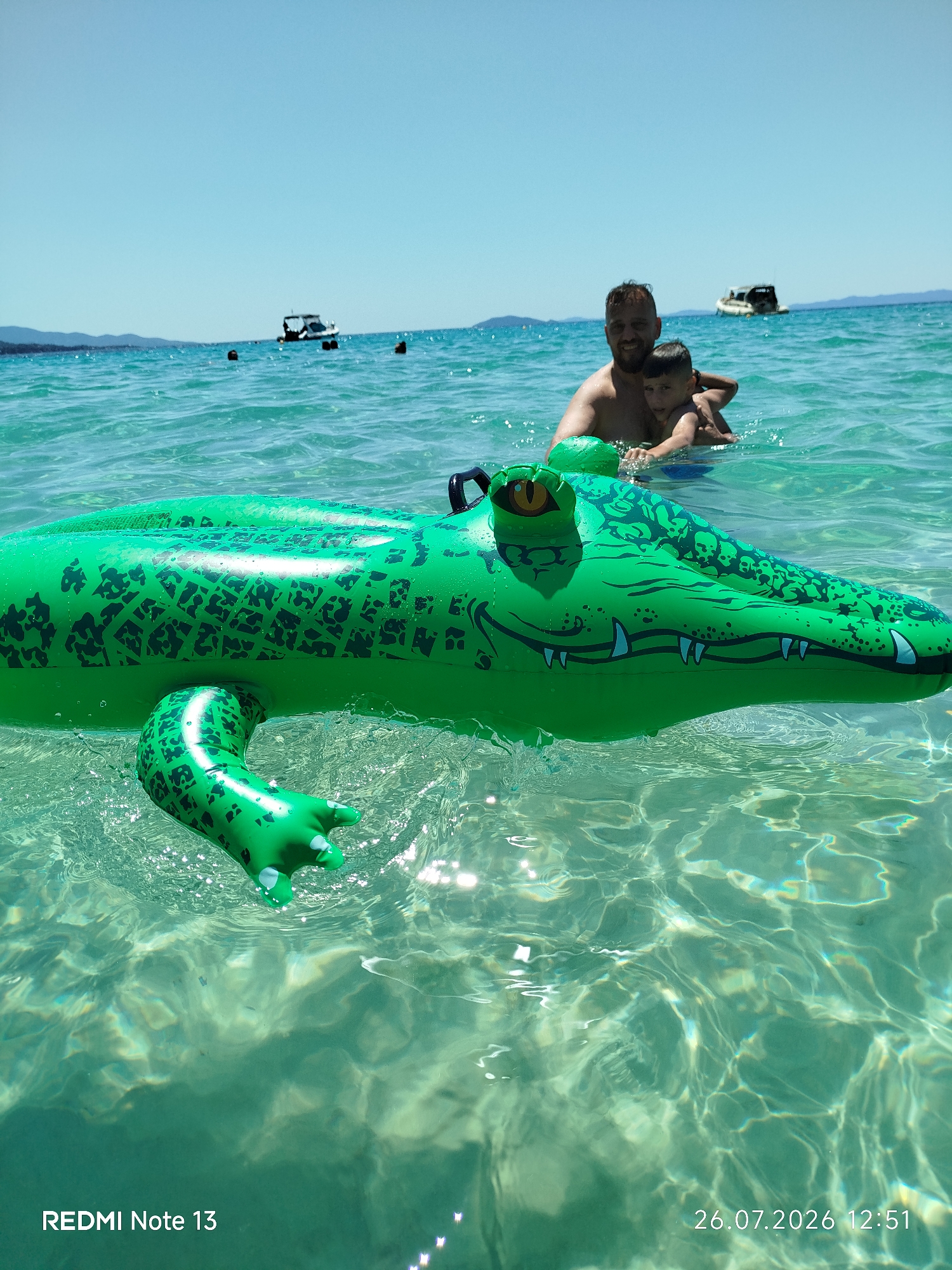A medium shot captures a man and a young boy in the clear, turquoise waters of Sithonia Municipal Unit, Greece. The man, with short dark hair and a beard, is holding the boy close. They are positioned behind a large, green inflatable alligator pool toy, which dominates the foreground. The alligator toy is partially submerged and appears to be floating. The water is transparent, allowing the sandy bottom and dappled sunlight to be visible. In the background, a few boats are moored on the horizon, and the coastline with hills is visible under a clear blue sky. The image is tagged with "REDMI Note 13" and a date and time stamp of "26.07.2026 12:51".