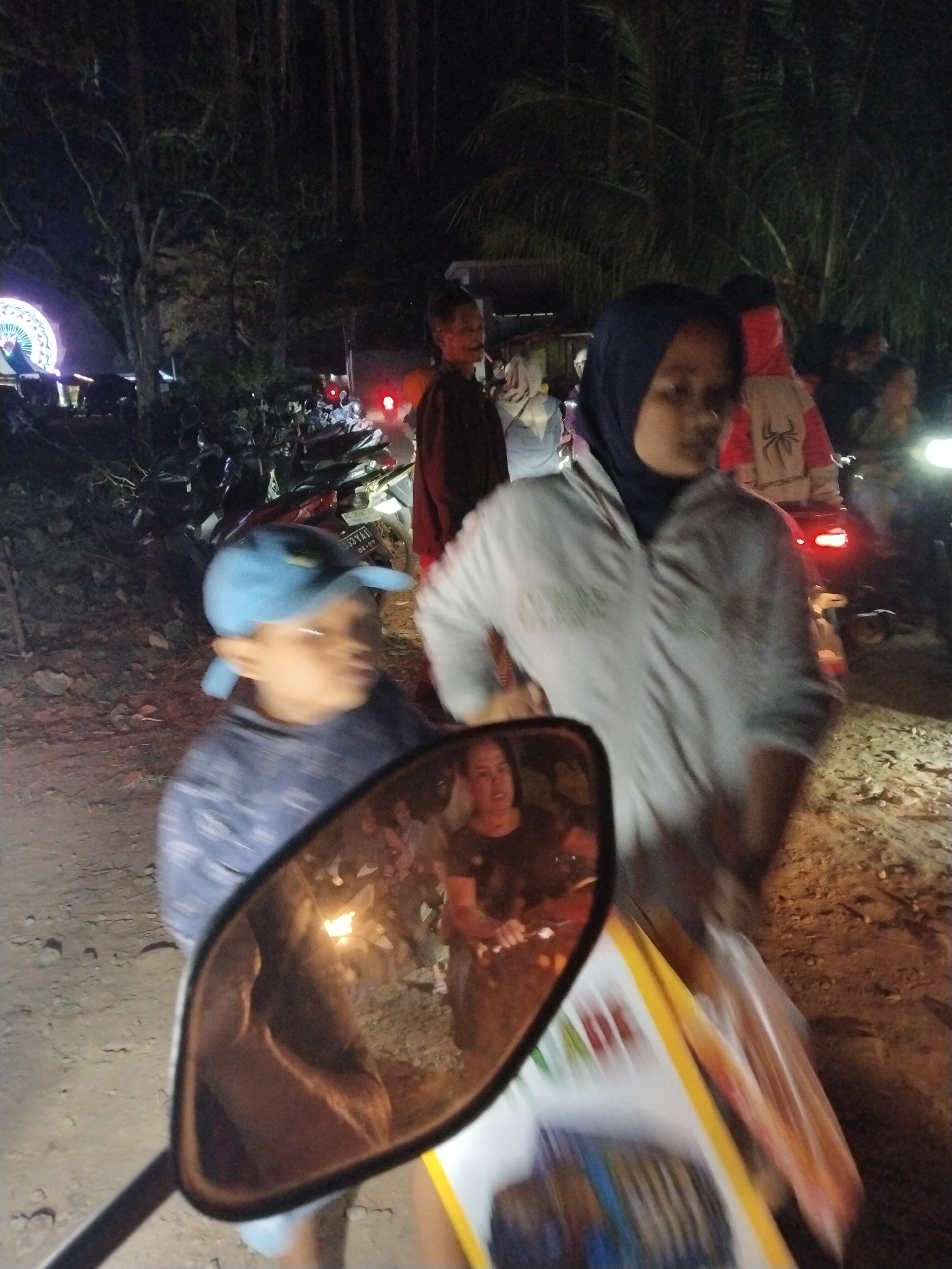 In Tasikmadu, Indonesia, at night, a young boy wearing a blue baseball cap and a blue patterned shirt is seen in the foreground. He is looking to his left, and his image is partially reflected in a motorcycle's rearview mirror. The mirror also reflects other people and lights. Behind the boy, a woman wearing a hijab and a light-colored jacket stands, holding a plastic bag. To the left of the boy, another person in a dark top is visible. In the background, numerous motorcycles are parked, and a Ferris wheel-like structure with lights can be seen in the distance. The ground appears to be dirt and gravel. The scene suggests a lively outdoor gathering or event at night in a public area.